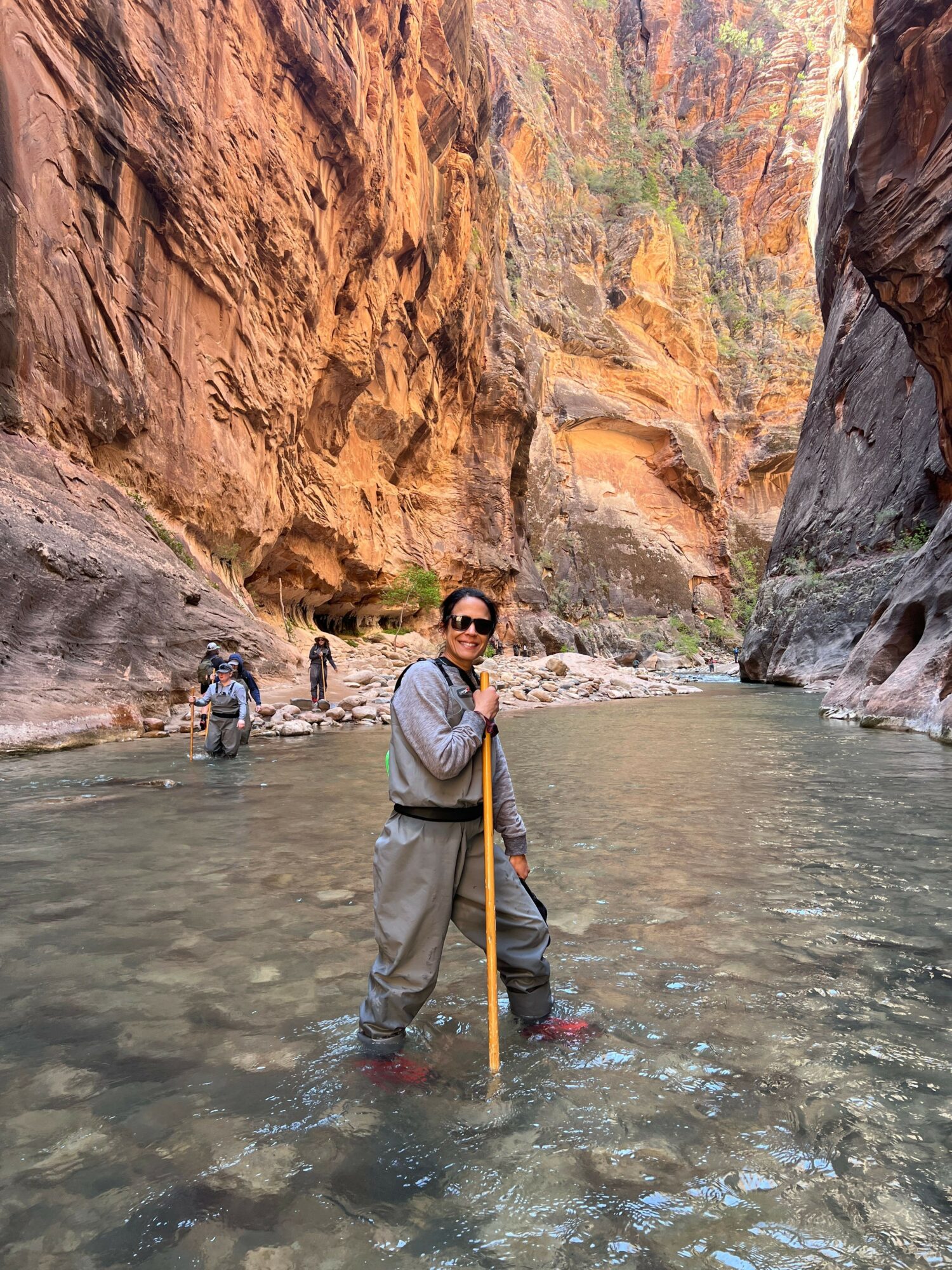 Person standing in river with canyon walls in background, holding a walking stick, wearing sunglasses and outdoor clothing.
