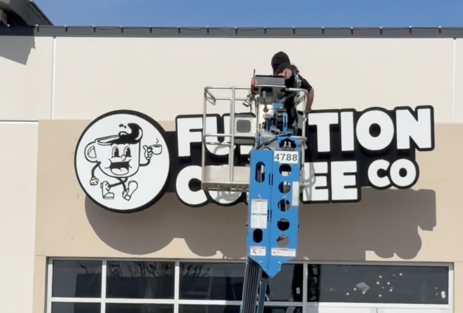 Worker on lift installing a sign on a building exterior, with a logo and text for a coffee company.