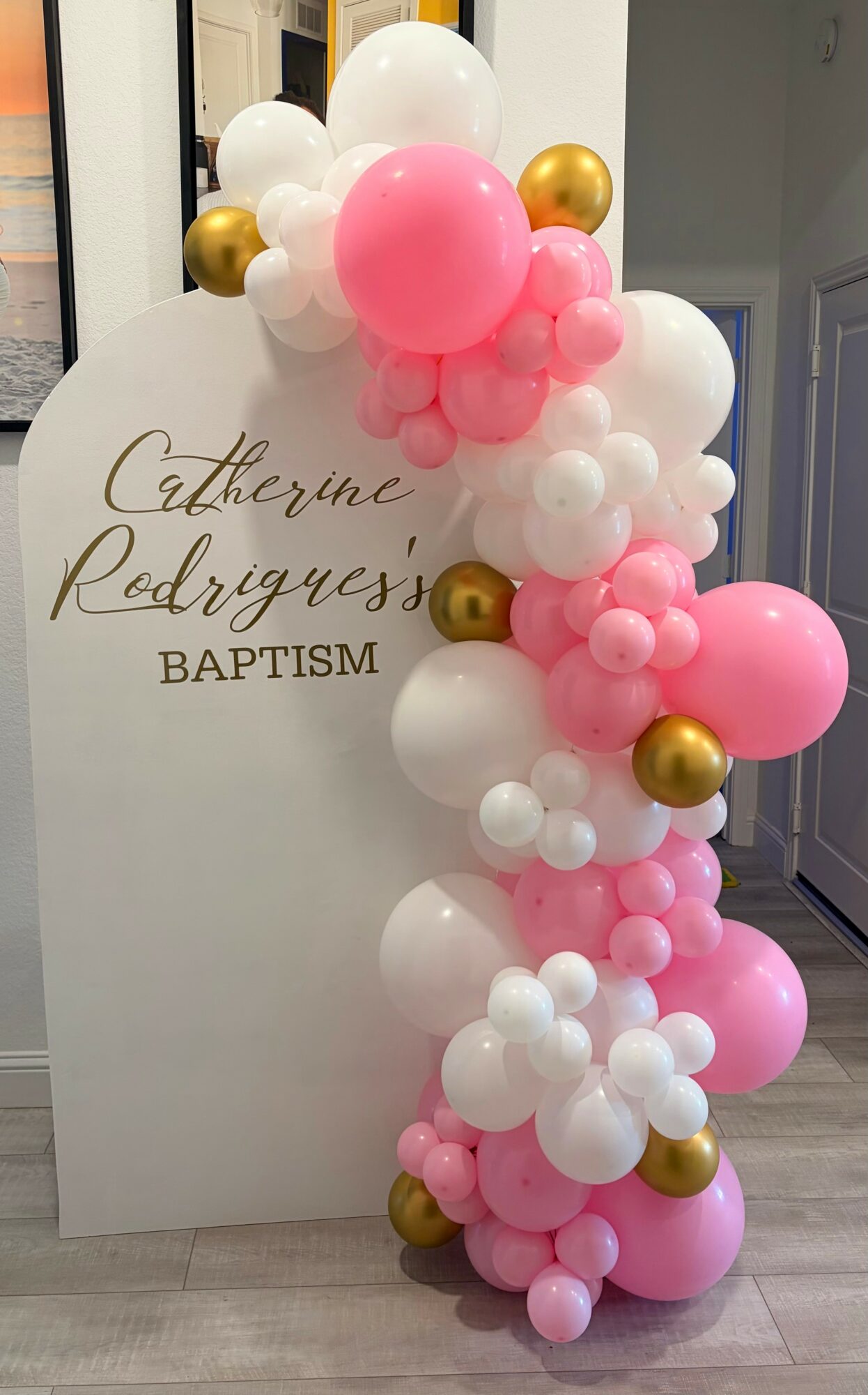 Balloon arrangement with pink, white, and gold balloons next to a baptism sign.