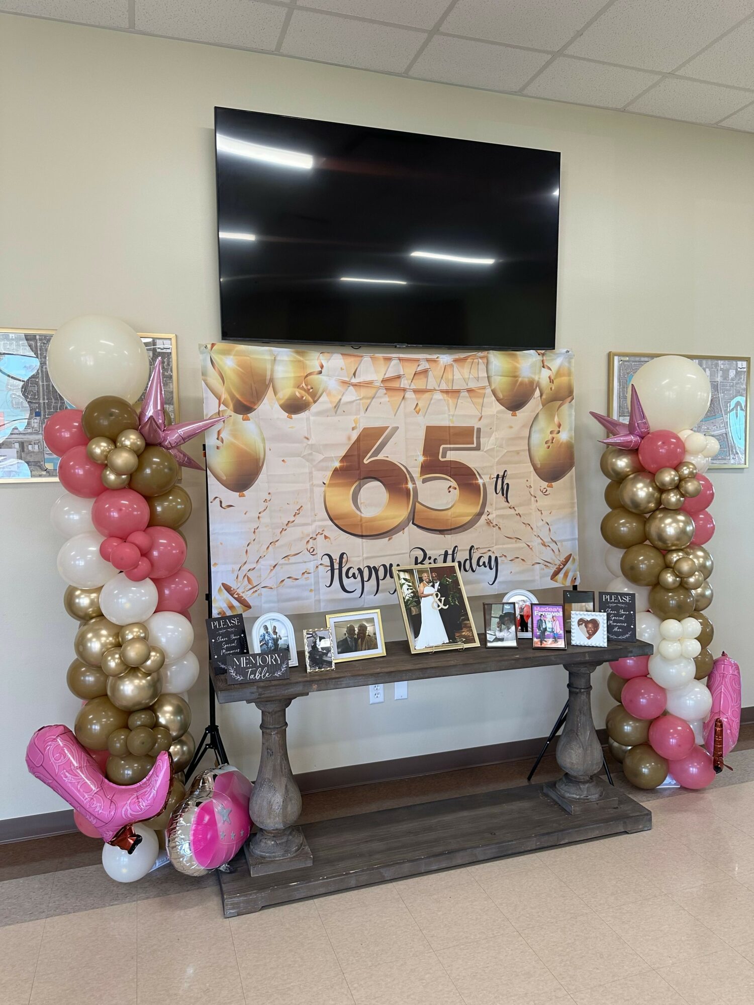 Decorative birthday display with balloons, photos, and a large '65' sign on a table, in front of a wall with a banner.