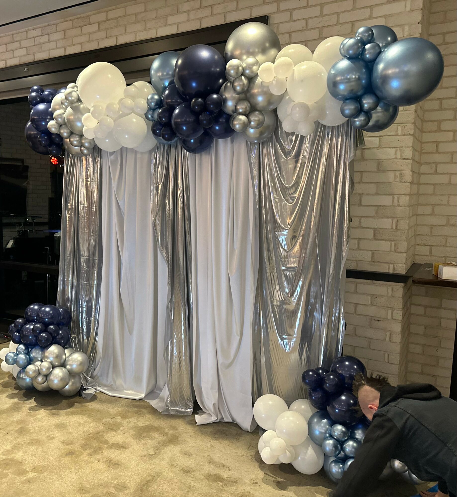Balloon arch with white, silver, and blue balloons over silver draped fabric backdrop, with a person crouching nearby.