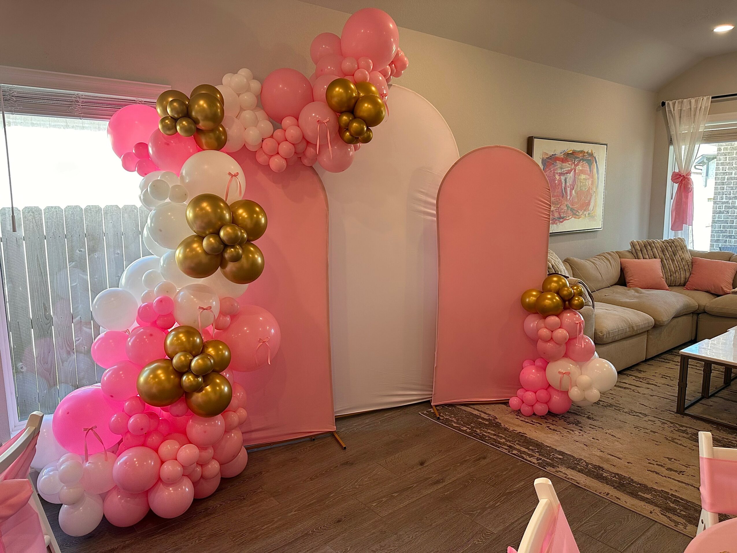 Decorative backdrop with pink, white, and gold balloons arranged around pink panels in a room with a sofa and window.