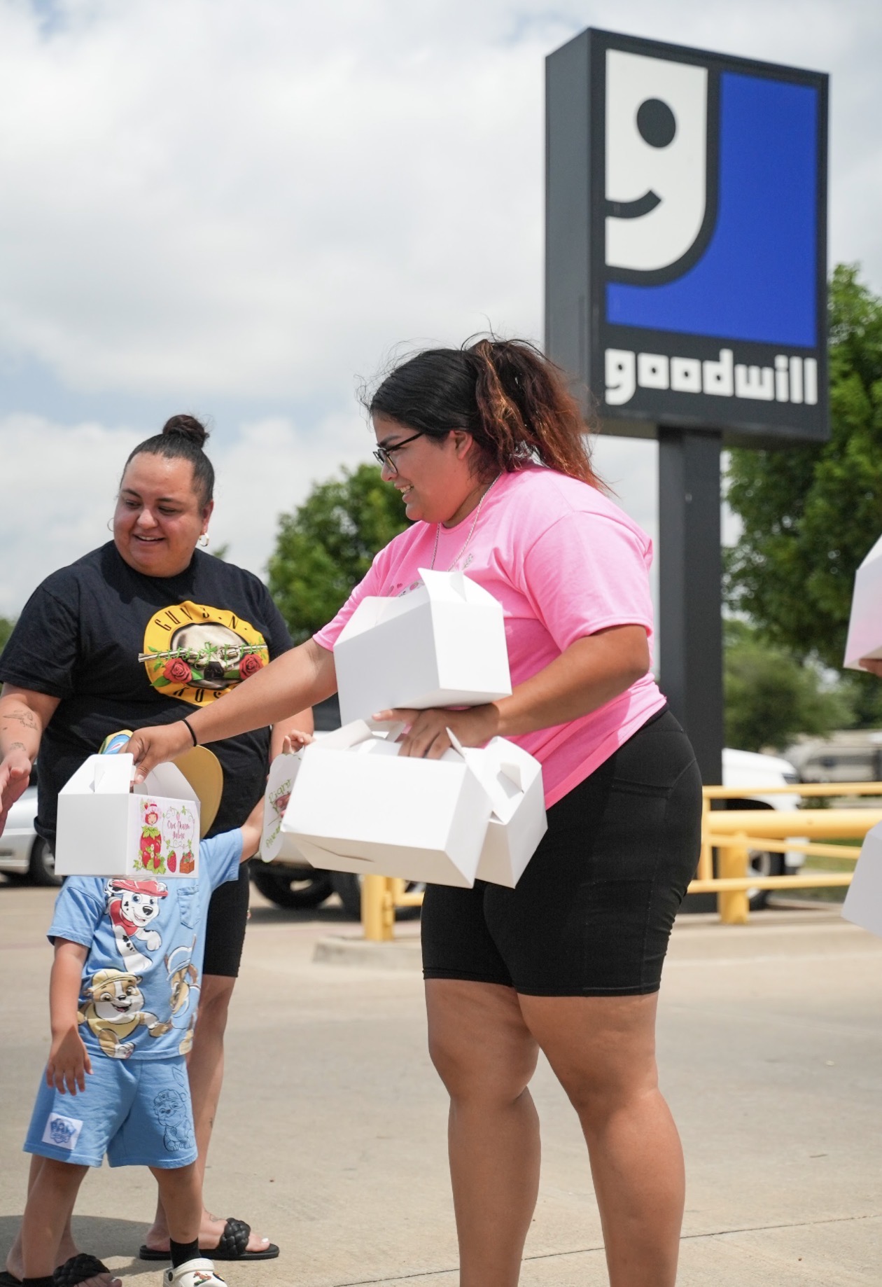 Two women exchanging food boxes outside a Goodwill store, one holding a cake, with a child nearby.