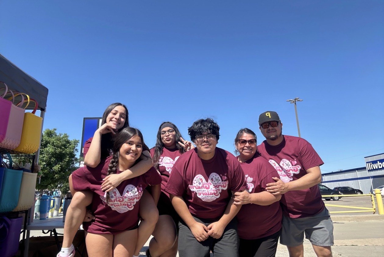 Group of six people outdoors on a sunny day, smiling and posing together, some with arms around each other.