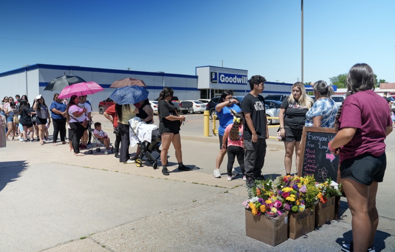 People standing in line outside a building with a Goodwill sign, some holding umbrellas, and a flower display nearby.