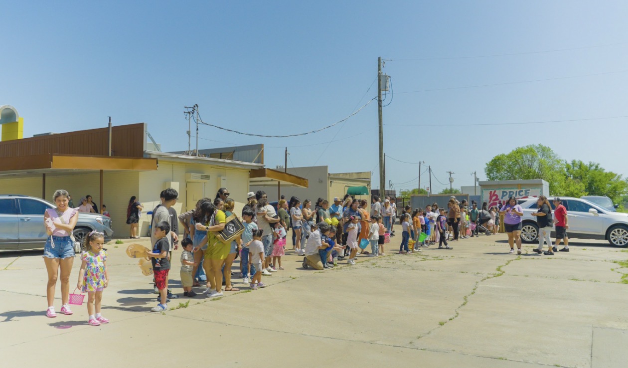 People standing in a line outdoors near a building and parked cars on a sunny day.