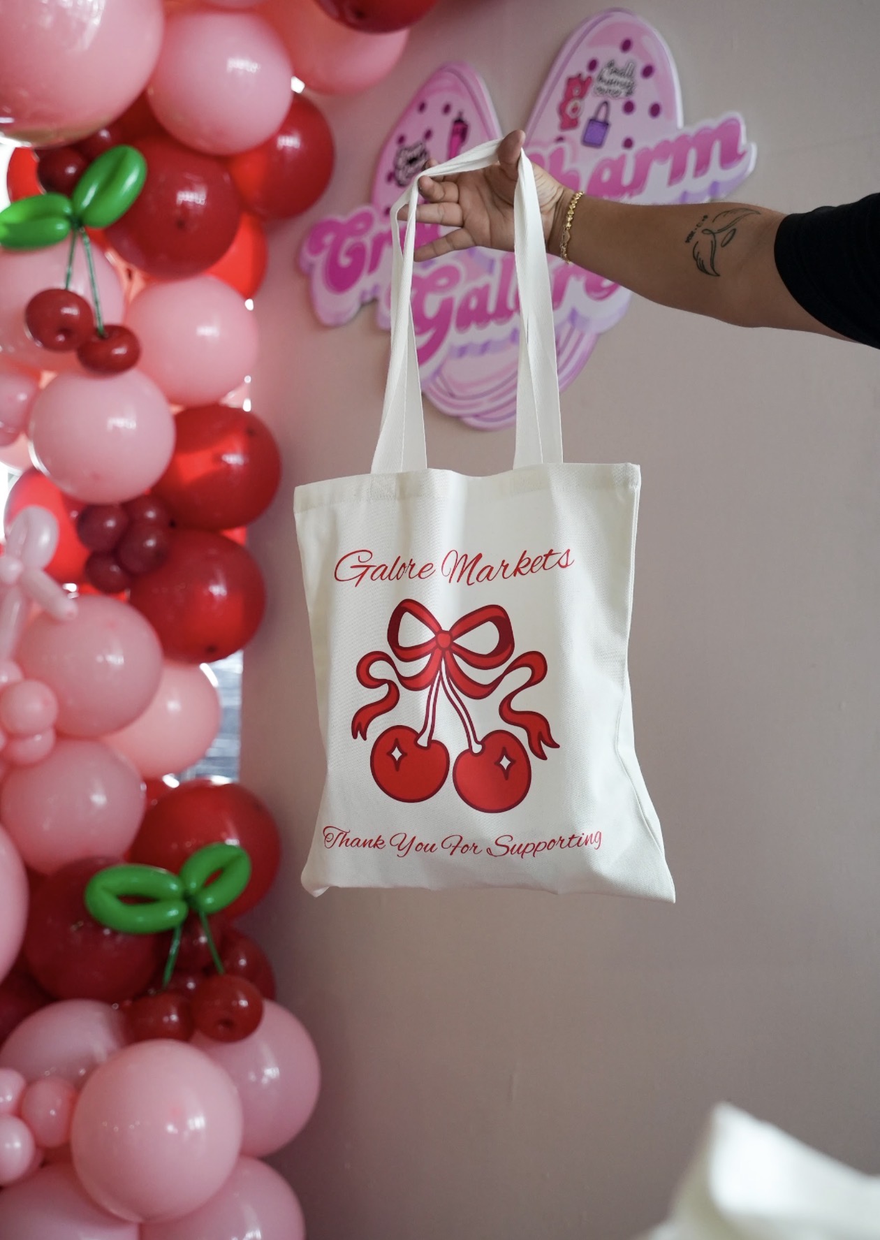 Person holding a tote bag with cherry graphic and text, decorated with pink and red balloons in the background.