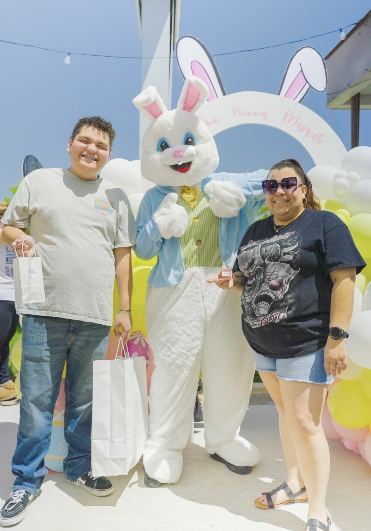 Two people stand with a person in a bunny costume at an outdoor event, smiling and holding shopping bags.