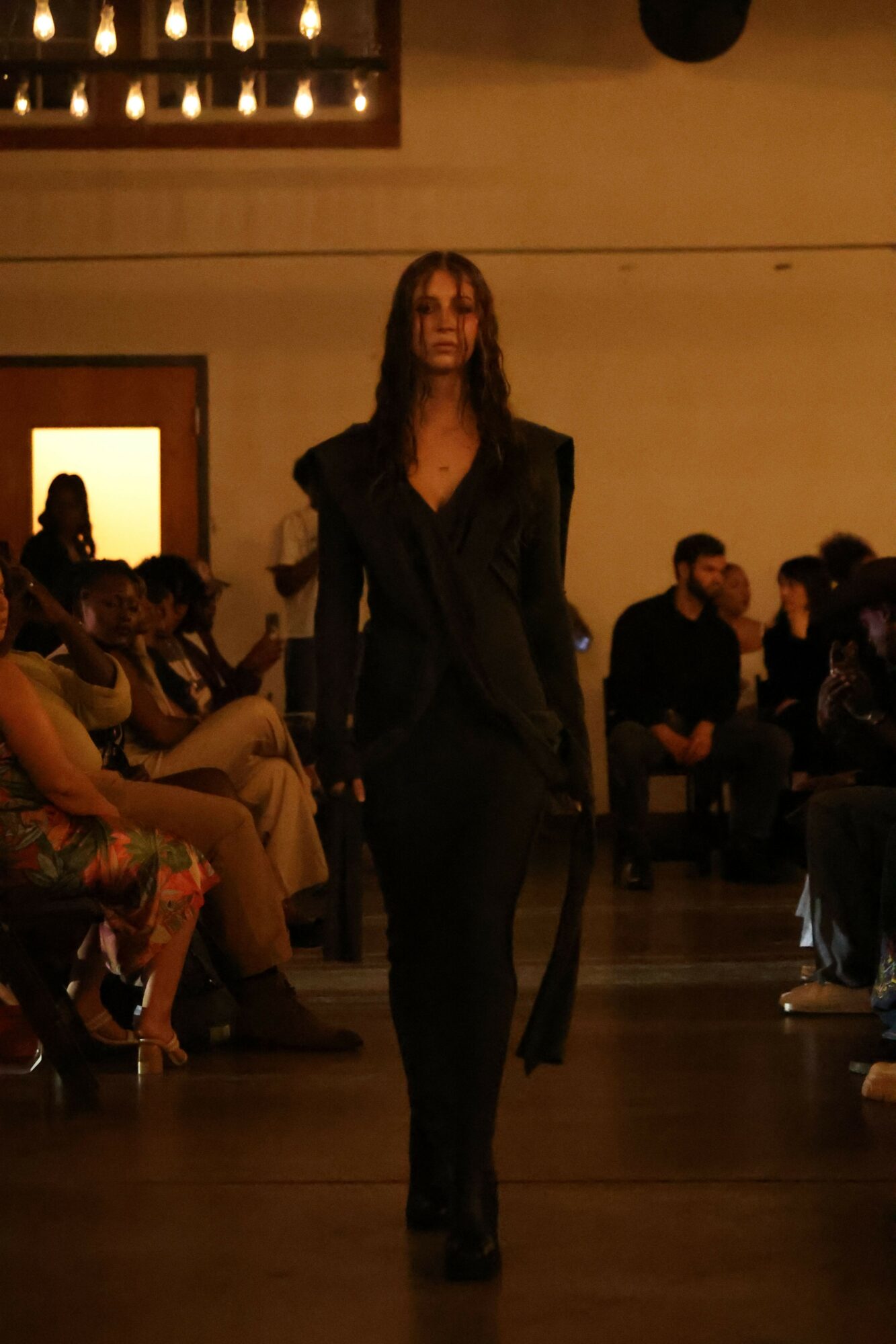 Model walking down runway in black outfit, audience seated on both sides, dim lighting in background.