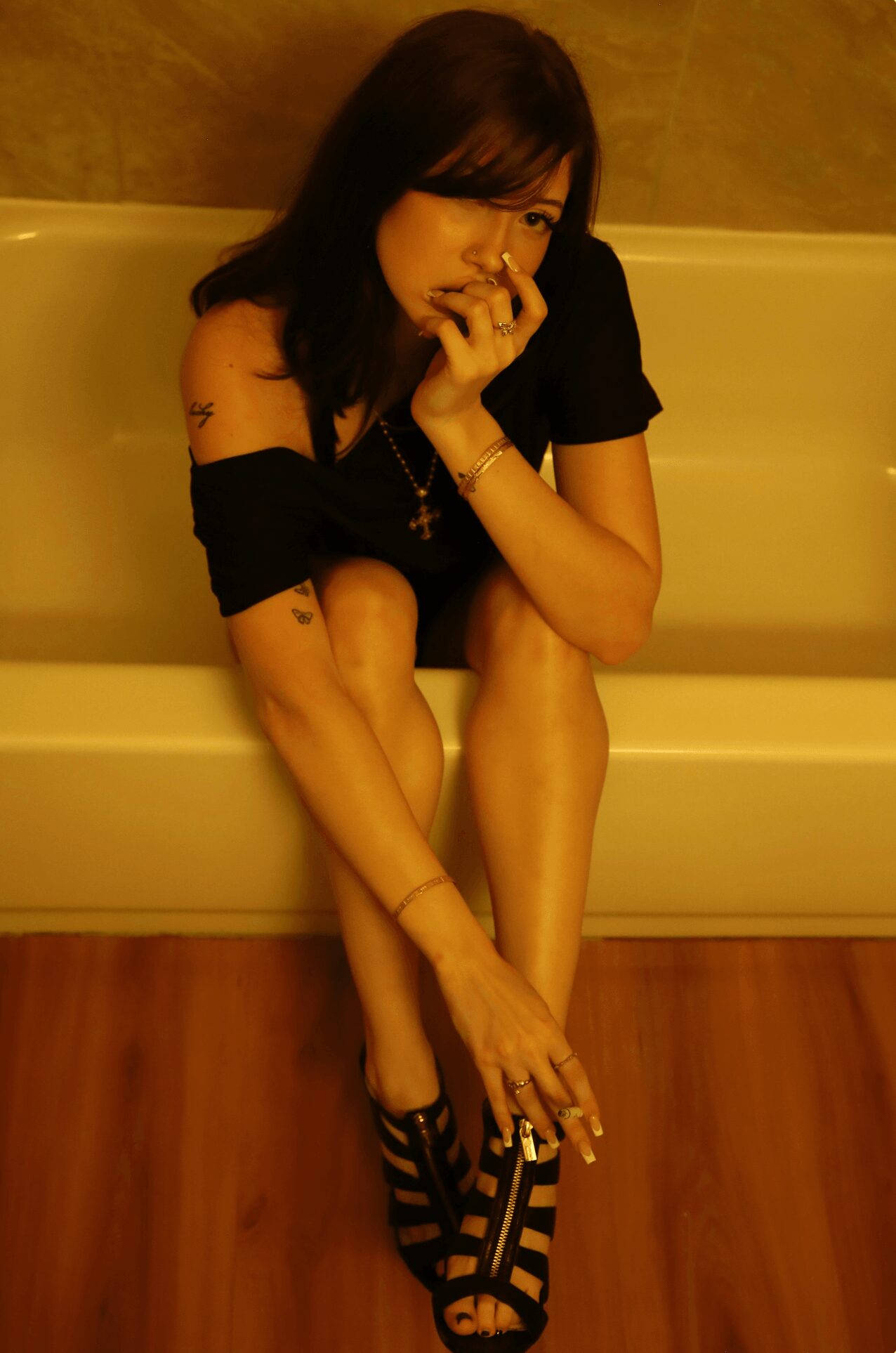 Woman sitting on a bench with legs crossed, wearing black dress and high heels, holding her face with one hand.