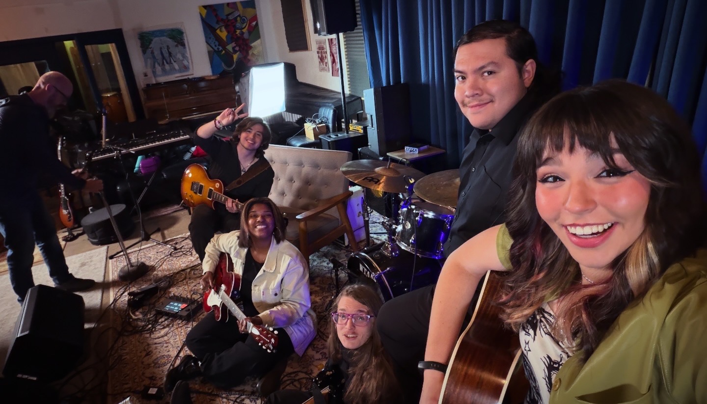 Group of people in a music studio, some playing instruments and others smiling at the camera.
