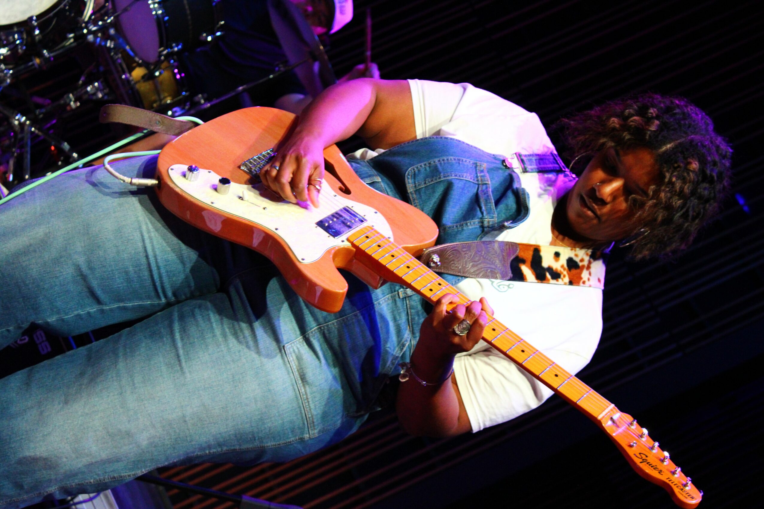 Person playing an electric guitar on stage with dark background, wearing a white shirt and jeans.