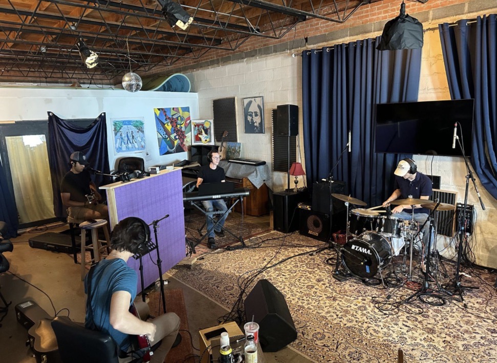 Music rehearsal space with musicians, instruments, and equipment, and a stage area with blue curtains and a large screen.