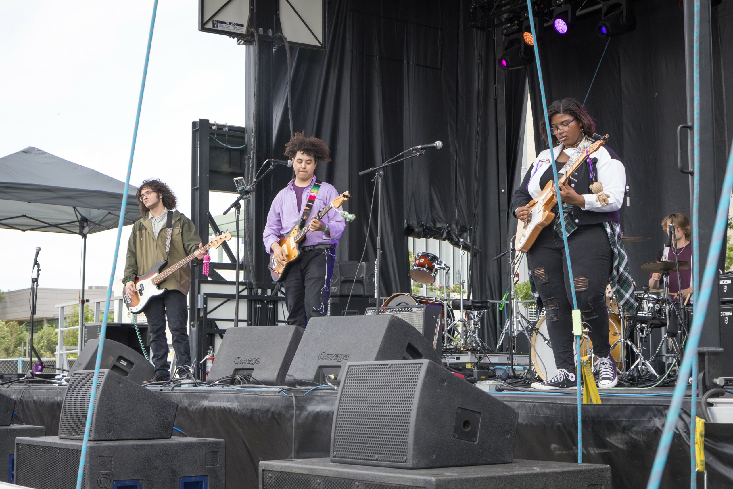 Four musicians perform on stage with guitars and microphones, black curtain backdrop, outdoor setting, sound equipment visible.