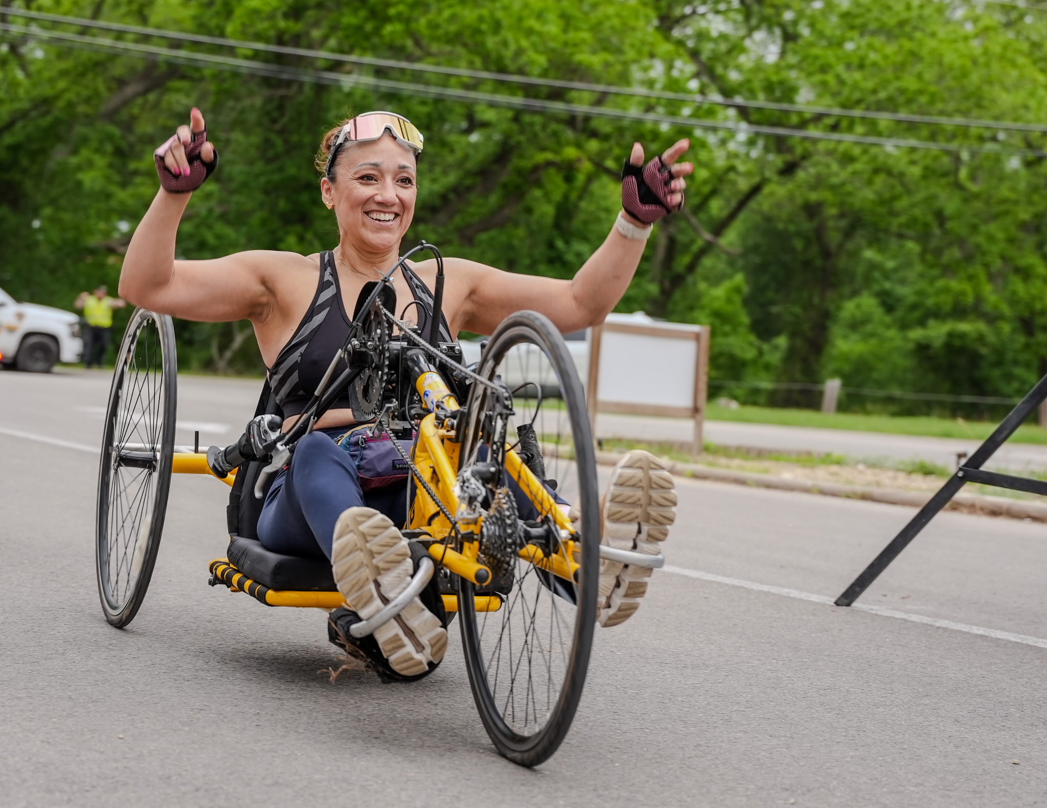 Person in a handcycle on the road, smiling and raising arms, with trees and a road in background.