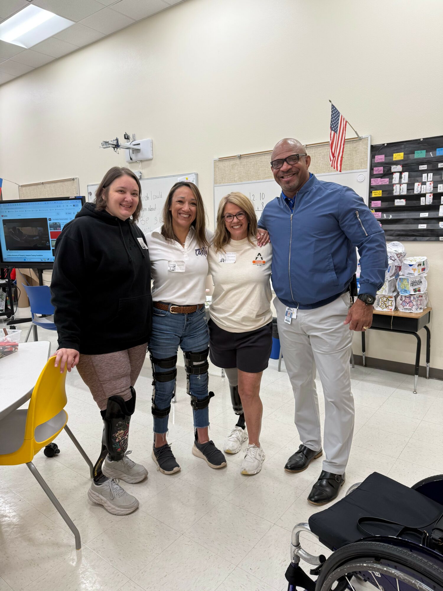 Four people standing together in a classroom, smiling, with a computer and American flag in the background.