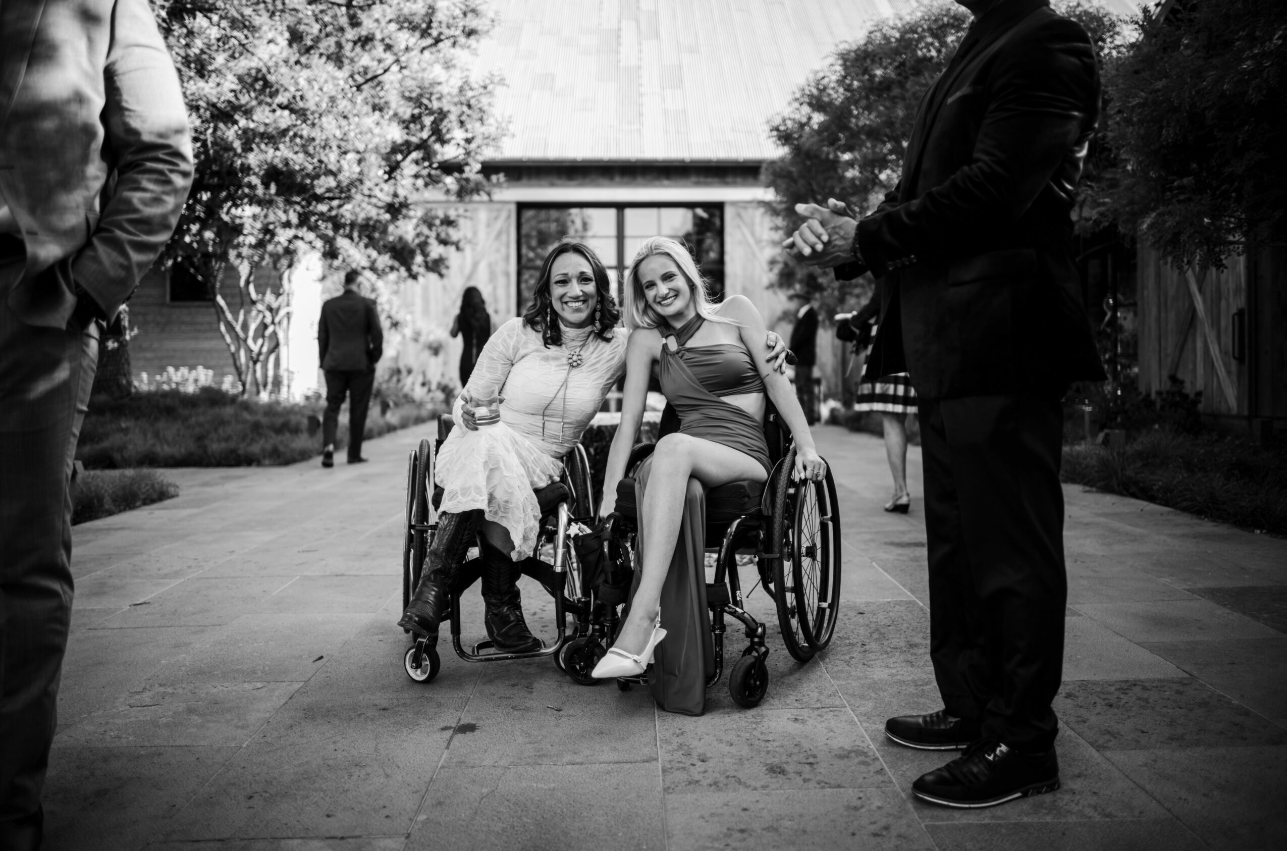 Two women in wheelchairs smiling, surrounded by people outdoors, with trees and a building in the background.