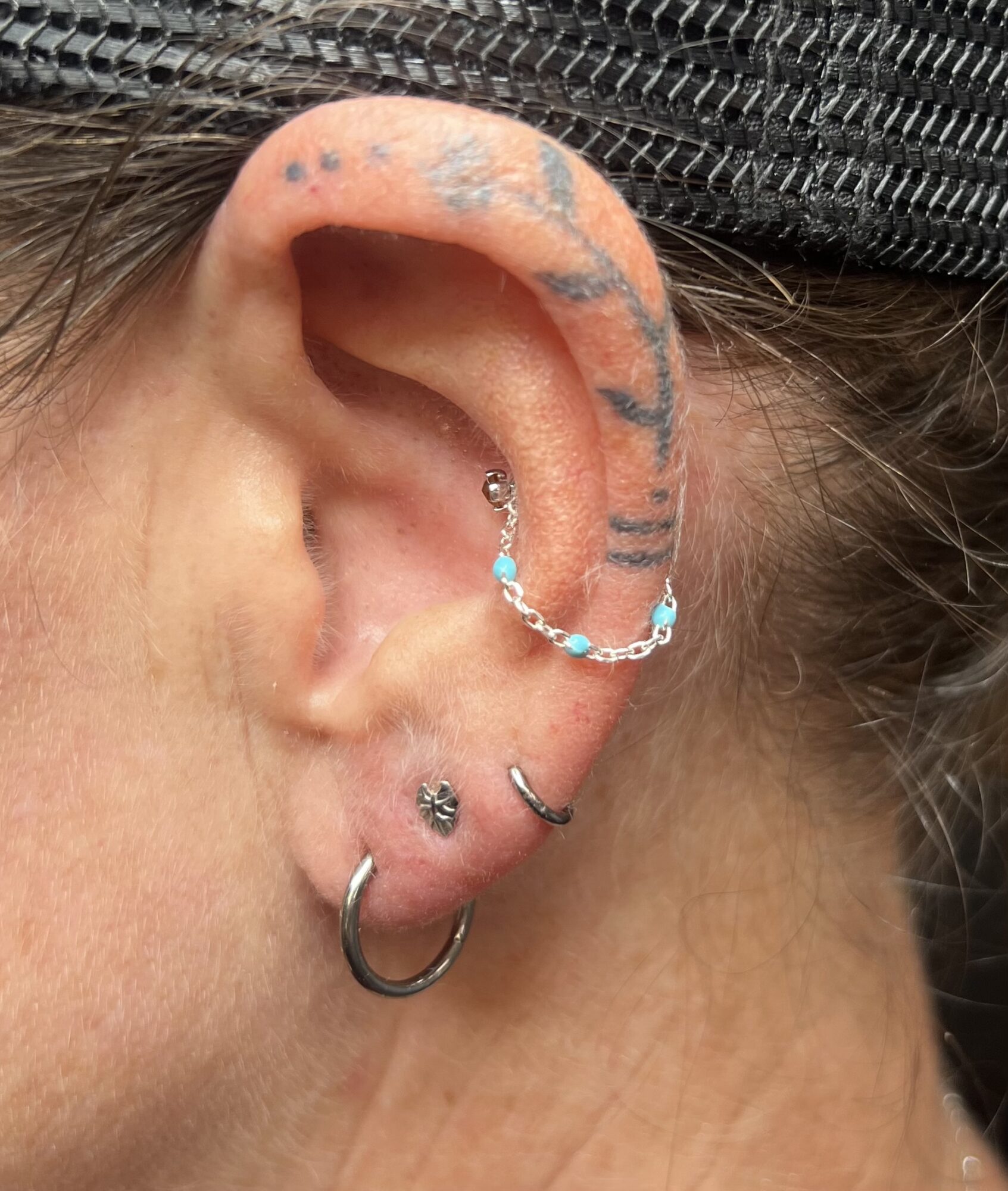Close-up of a person's ear with multiple piercings, tattoos, and jewelry, including a helix piercing with a chain and a hoop.