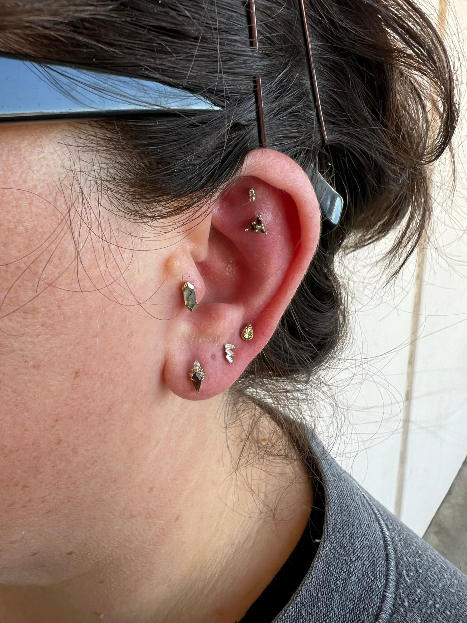 Close-up of a person's ear with multiple piercings and earrings, hair partially covering the ear, wearing glasses.