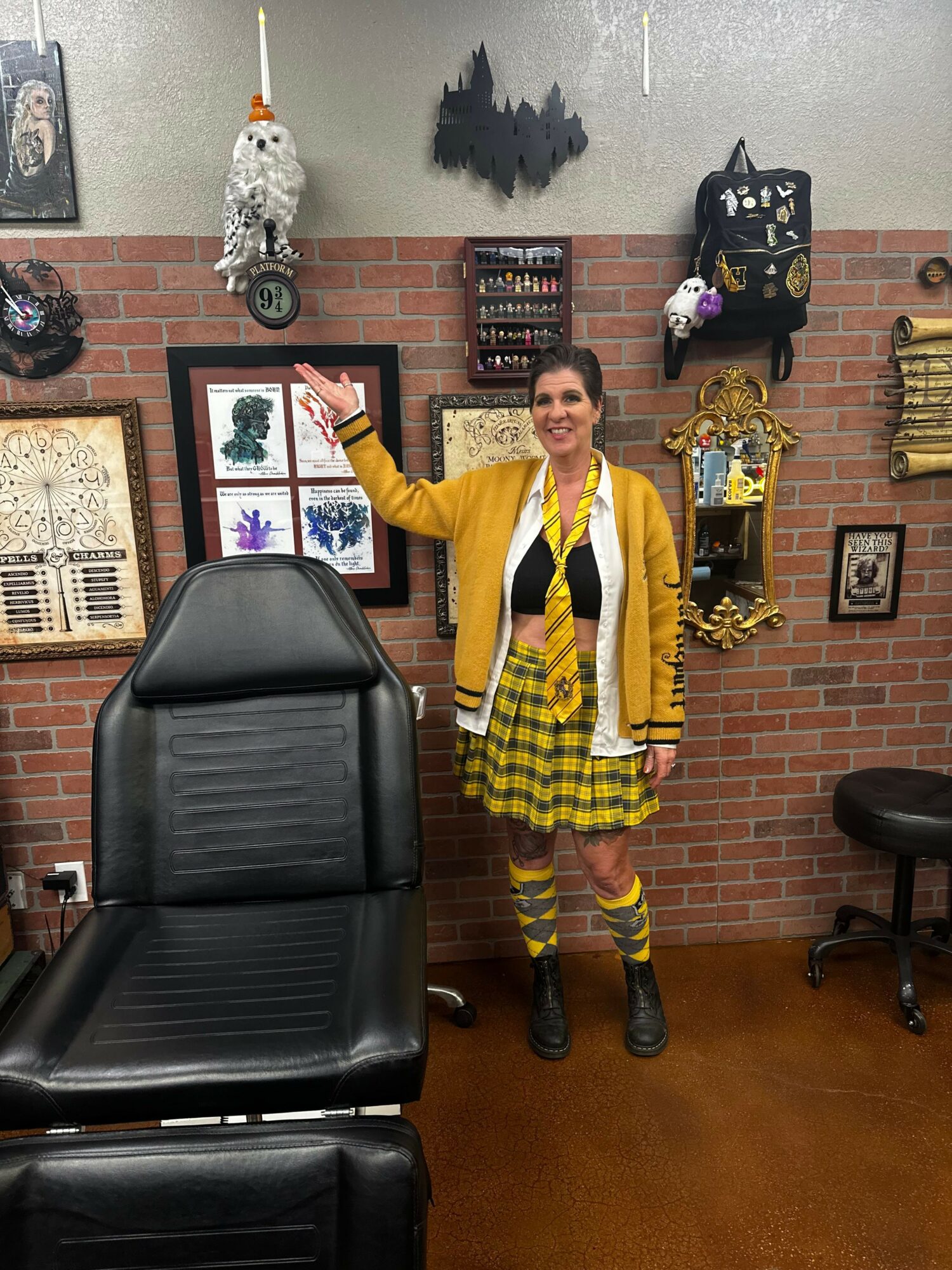 Woman standing in front of a brick wall decorated with framed art, shelves, and hanging ornaments, smiling and pointing upward.