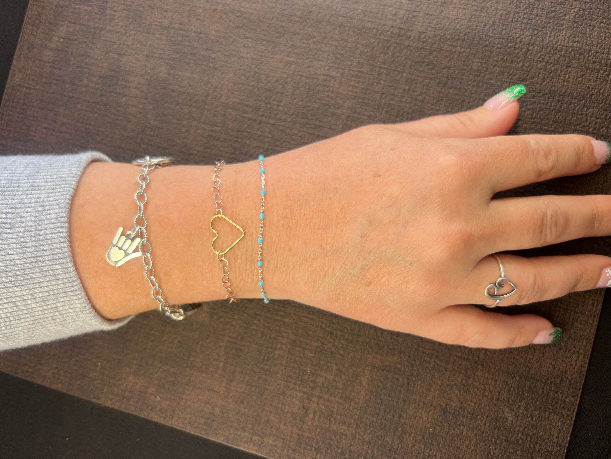 Hand resting on a surface with three bracelets and a ring, nails painted green, gray sleeve visible.
