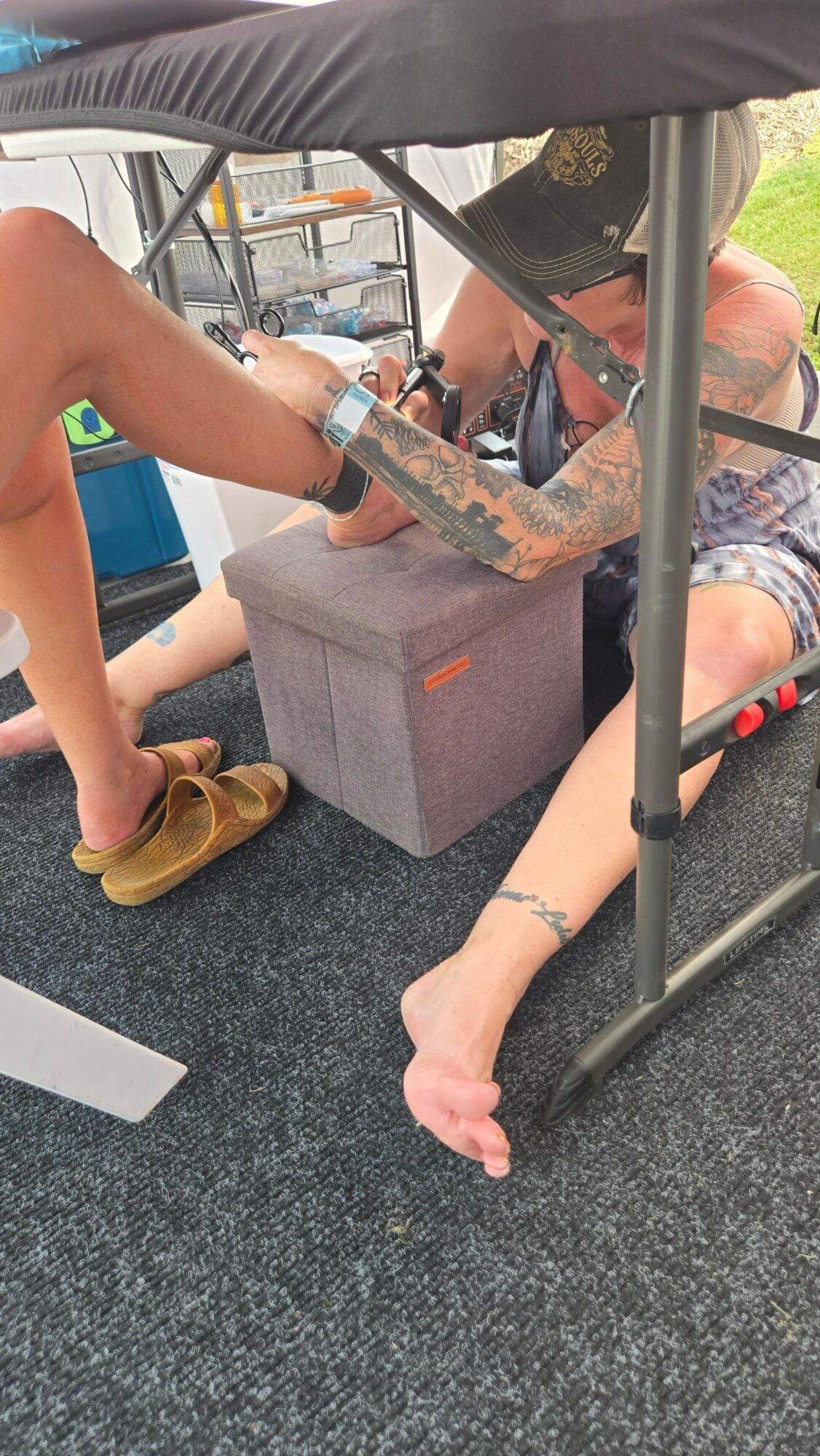 Person lying on ground with arm extended, receiving a tattoo from another person. They are under a canopy outdoors.