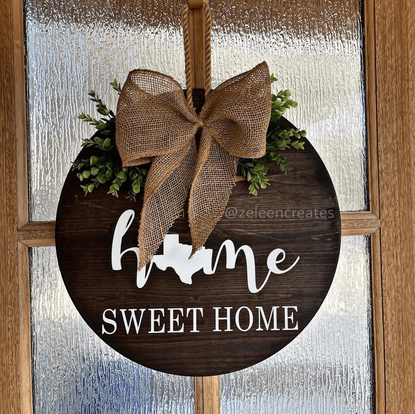 Round wooden sign with a bow and greenery, displaying 'home' and 'SWEET HOME' text, hanging on a door.