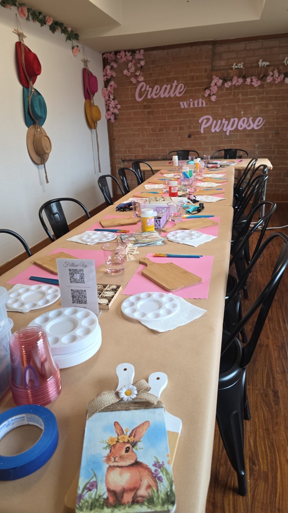 Long table set with pink tablecloth, plates, cups, and decorations, in a room with colorful wall art and a brick wall.