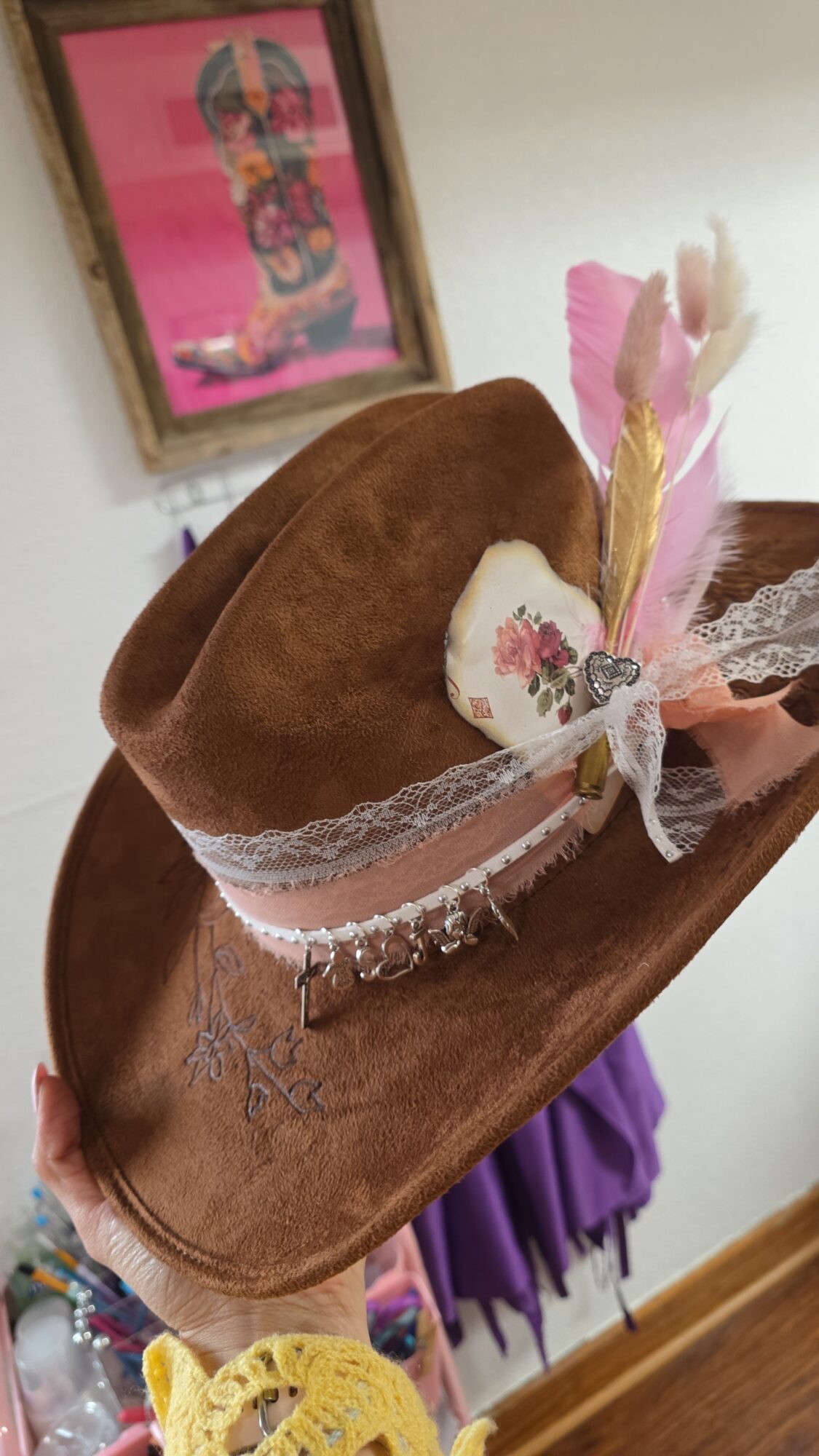 Brown hat with pink feathers, lace, and a decorative bow, placed on a wooden surface.