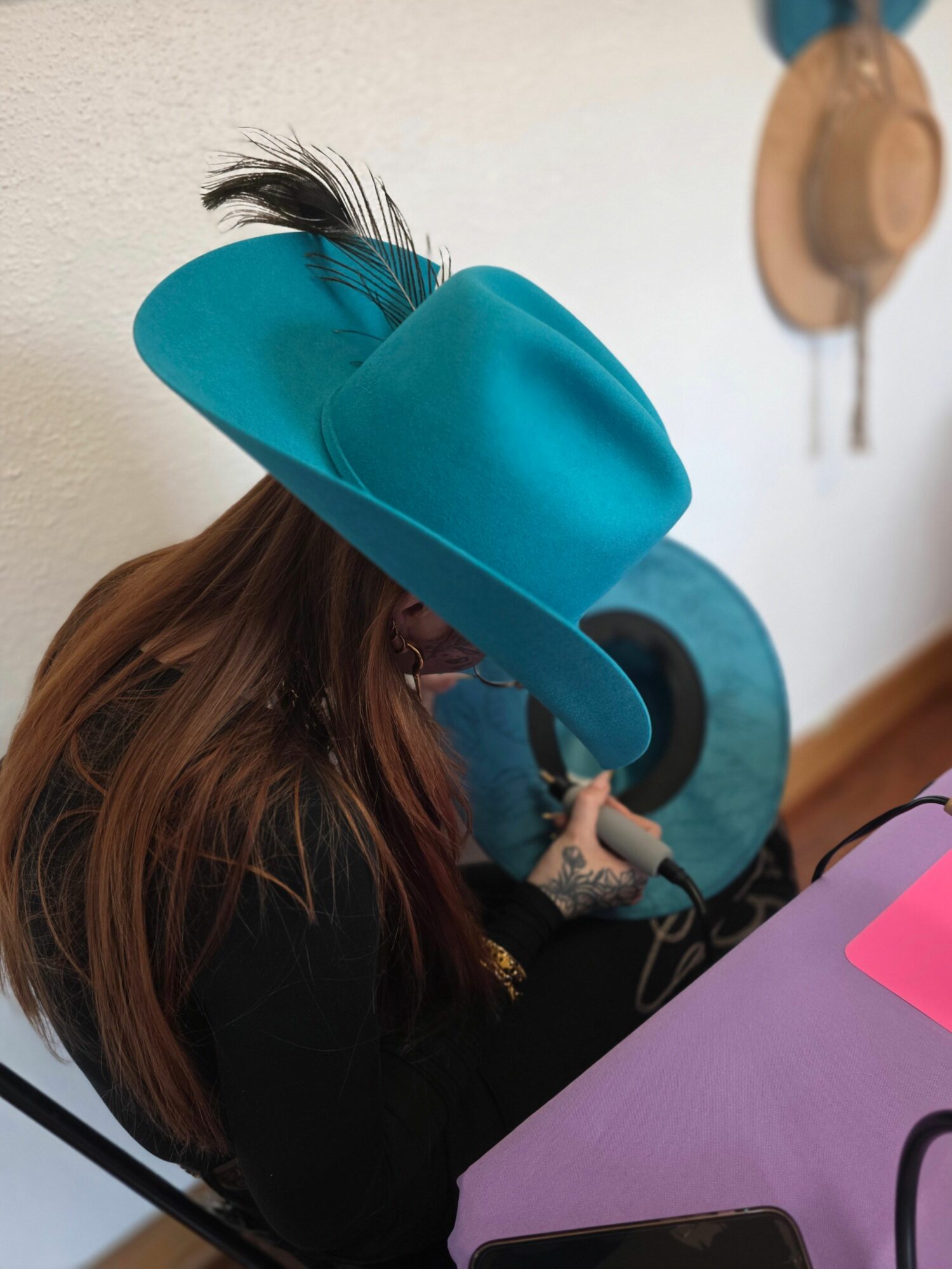 Person wearing a large turquoise hat with a feather, sitting at a table with a pink paper and a purple surface.