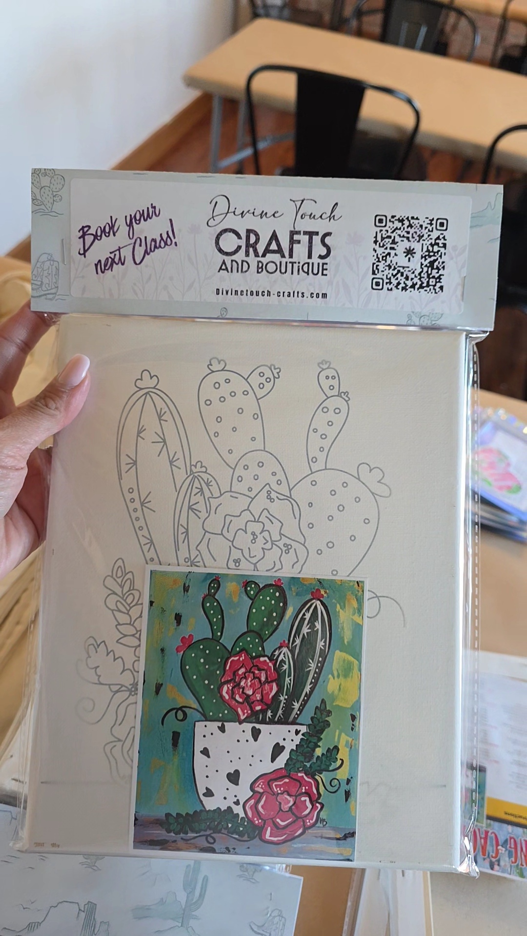 Hand holding craft kit with cactus and flower illustrations, in a room with tables and chairs.