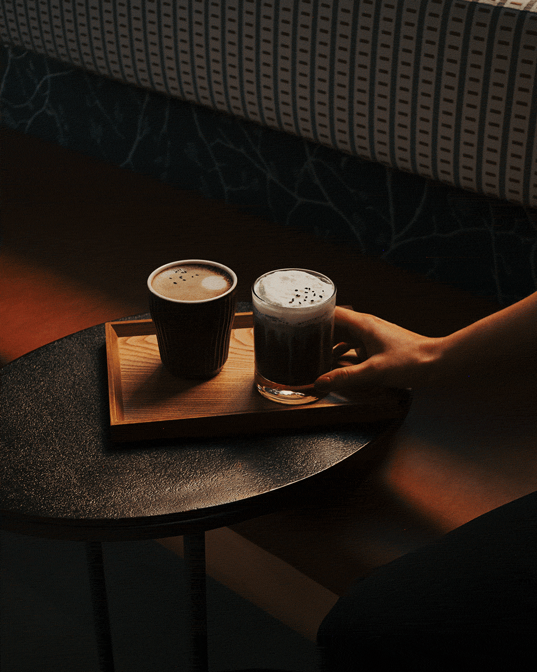 Two glasses of dark beer on a wooden tray, one person reaching for the glass, sitting on a dark surface.