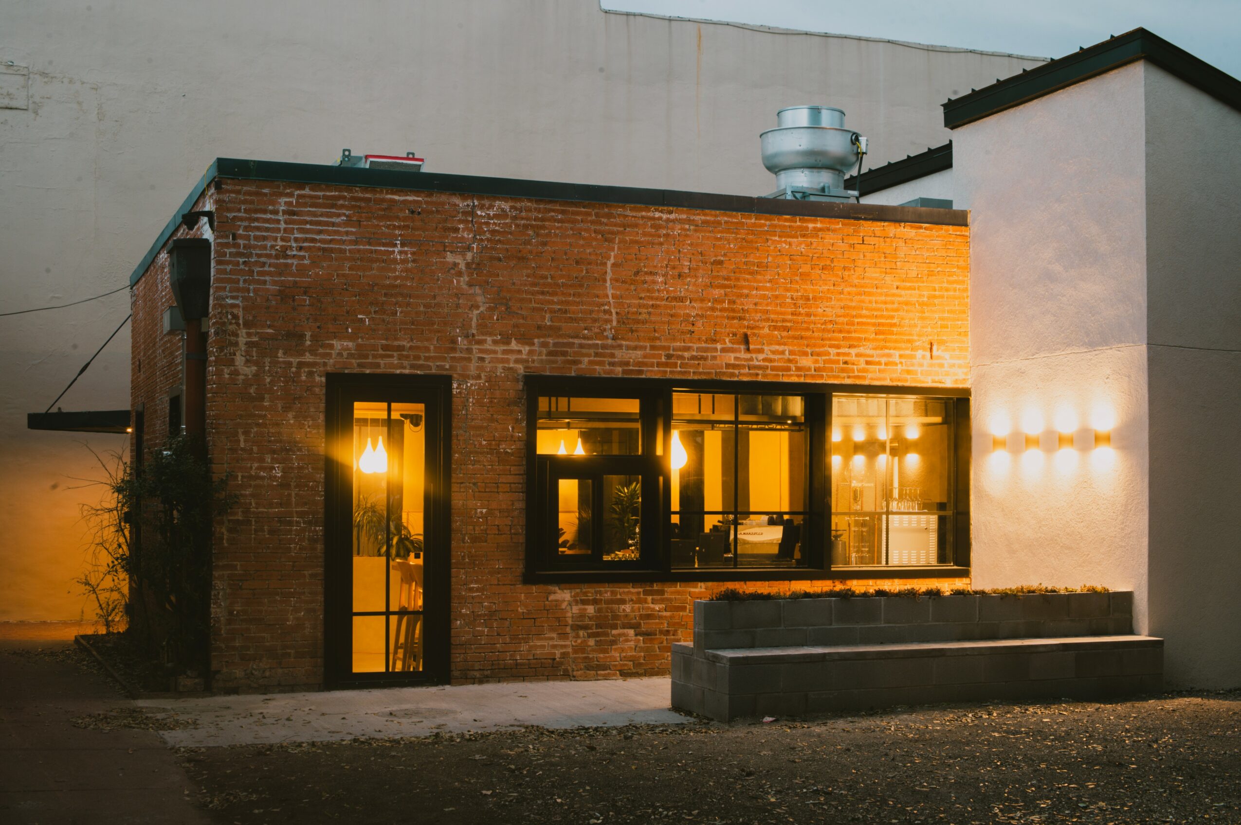 Brick building with large windows illuminated from inside, outdoor lighting, and a small concrete step in front.