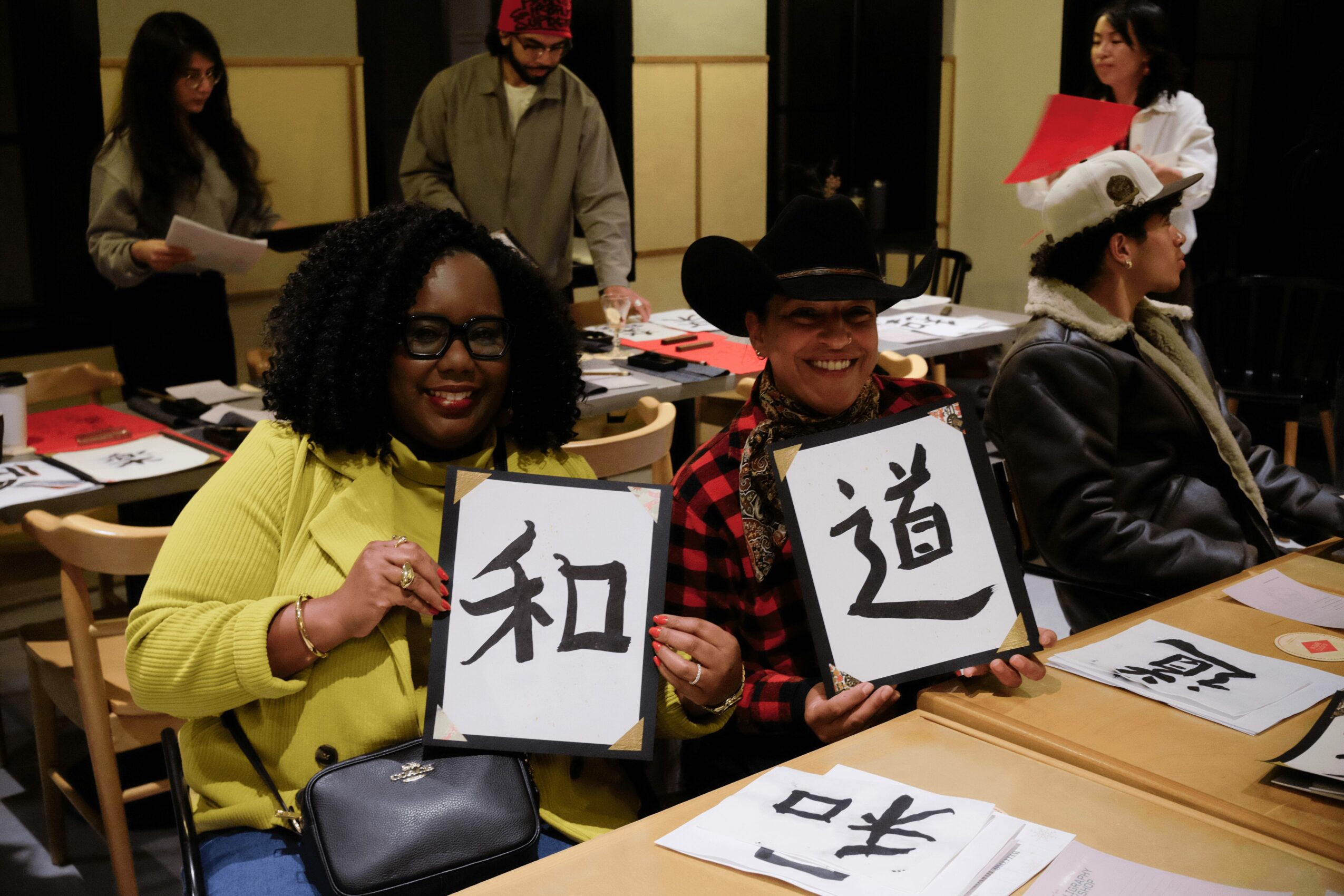 Two people sitting at a table holding calligraphy signs, smiling, in a room with others working on calligraphy.