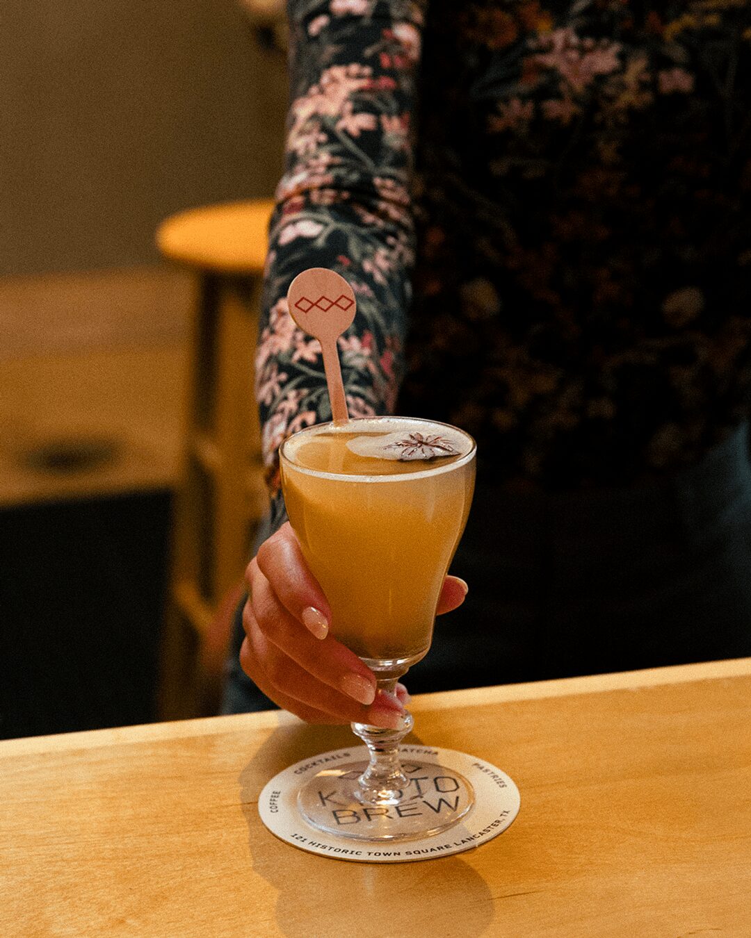 Person holding a cocktail glass with a yellow drink and a pink stirrer, standing at a wooden table.
