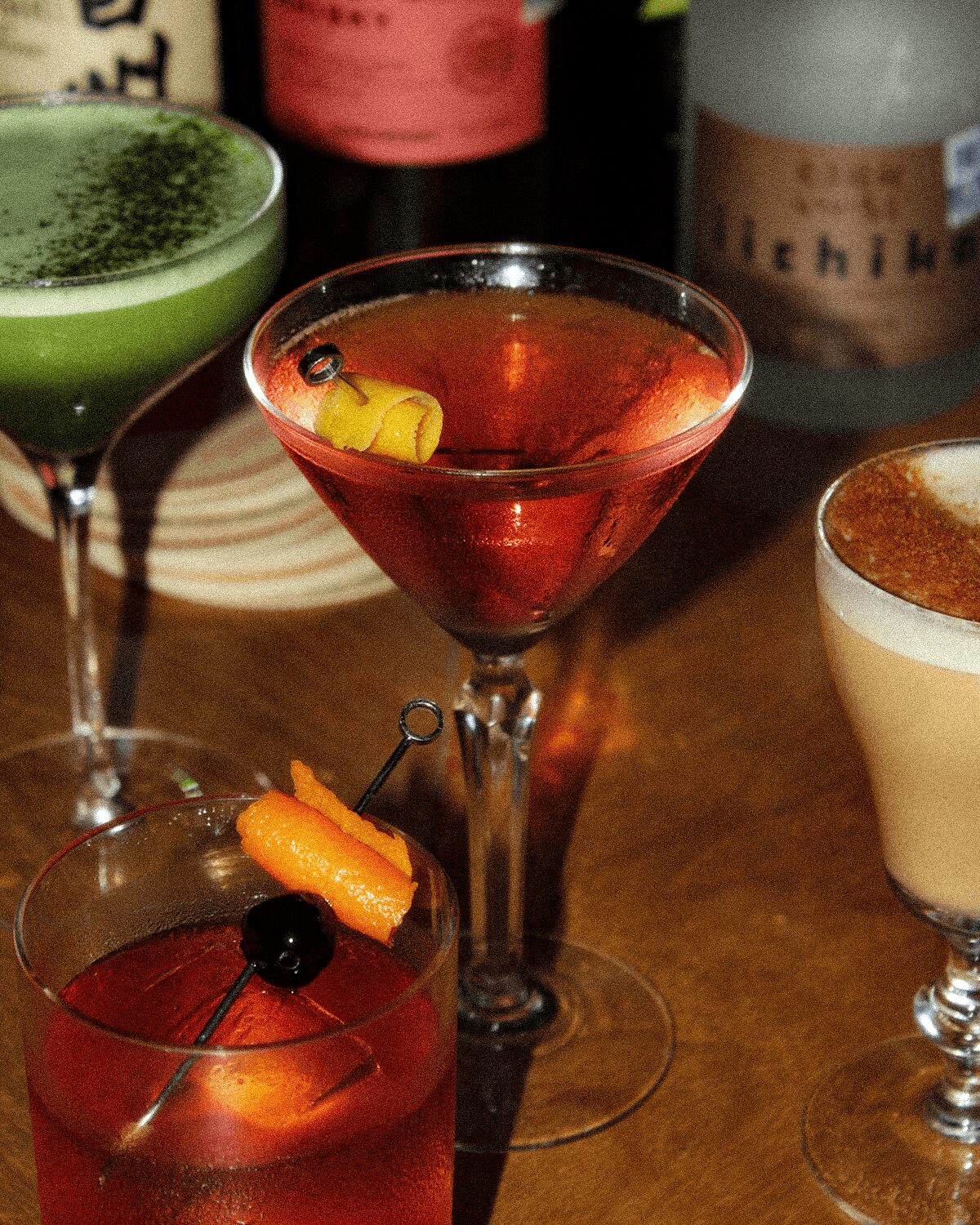 Three cocktails on a wooden table with bottles in the background, one garnished with a lemon twist and cherry.