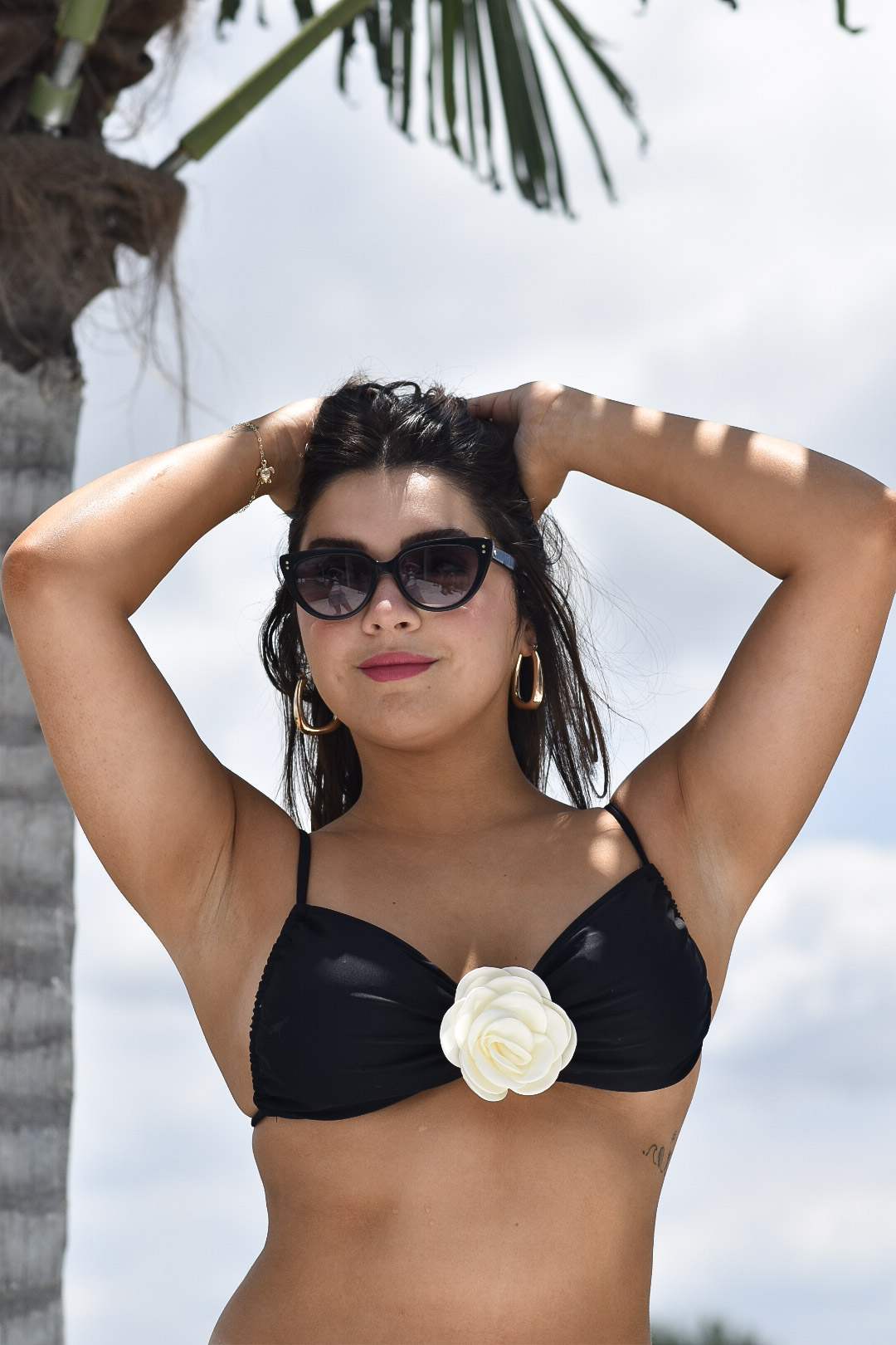 Woman in sunglasses and a black bikini top with a white flower, standing outdoors with a palm tree and sky.