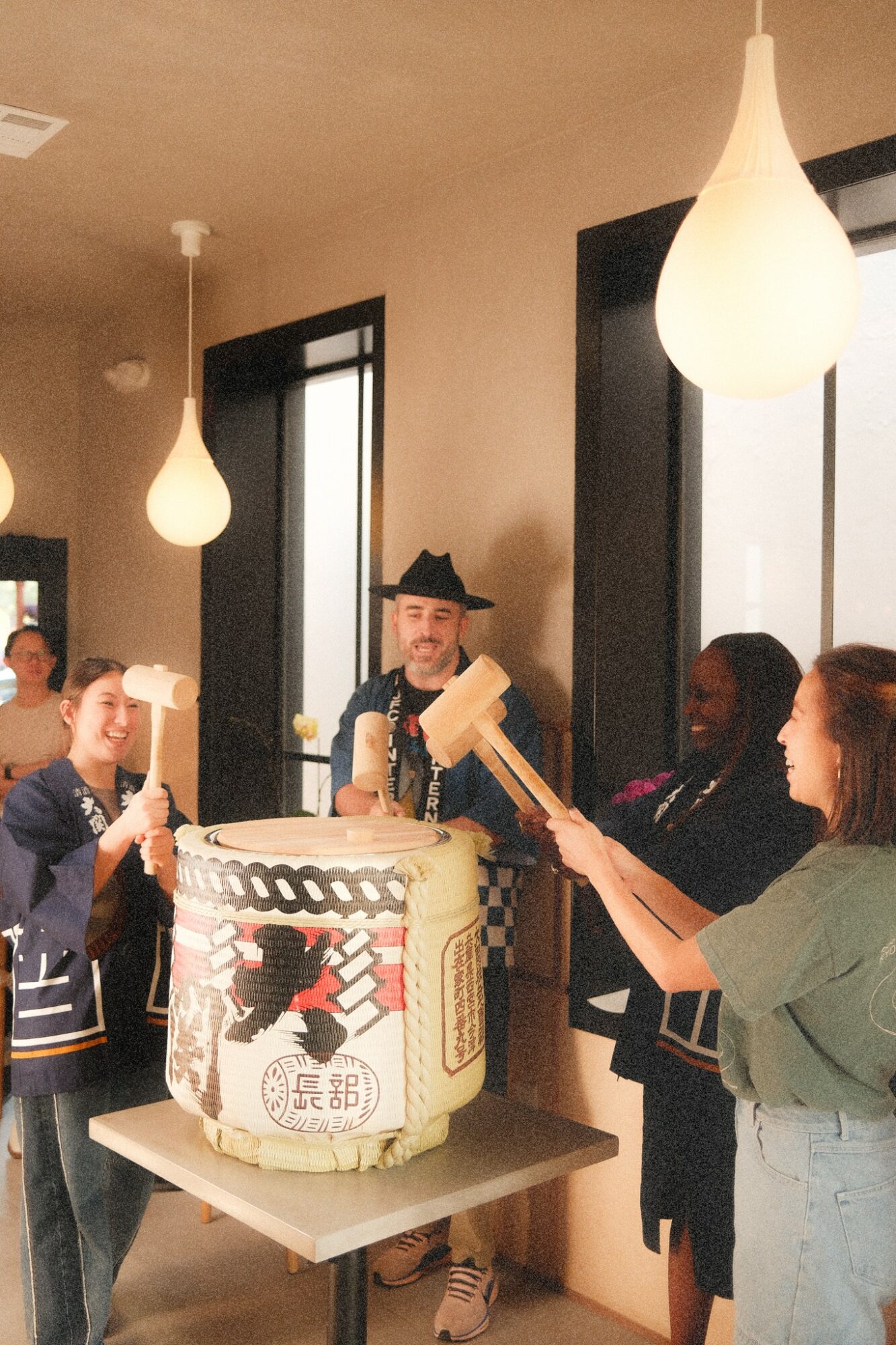 Group of people celebrating around a cake with a black and red design, in a room with hanging lights and windows.