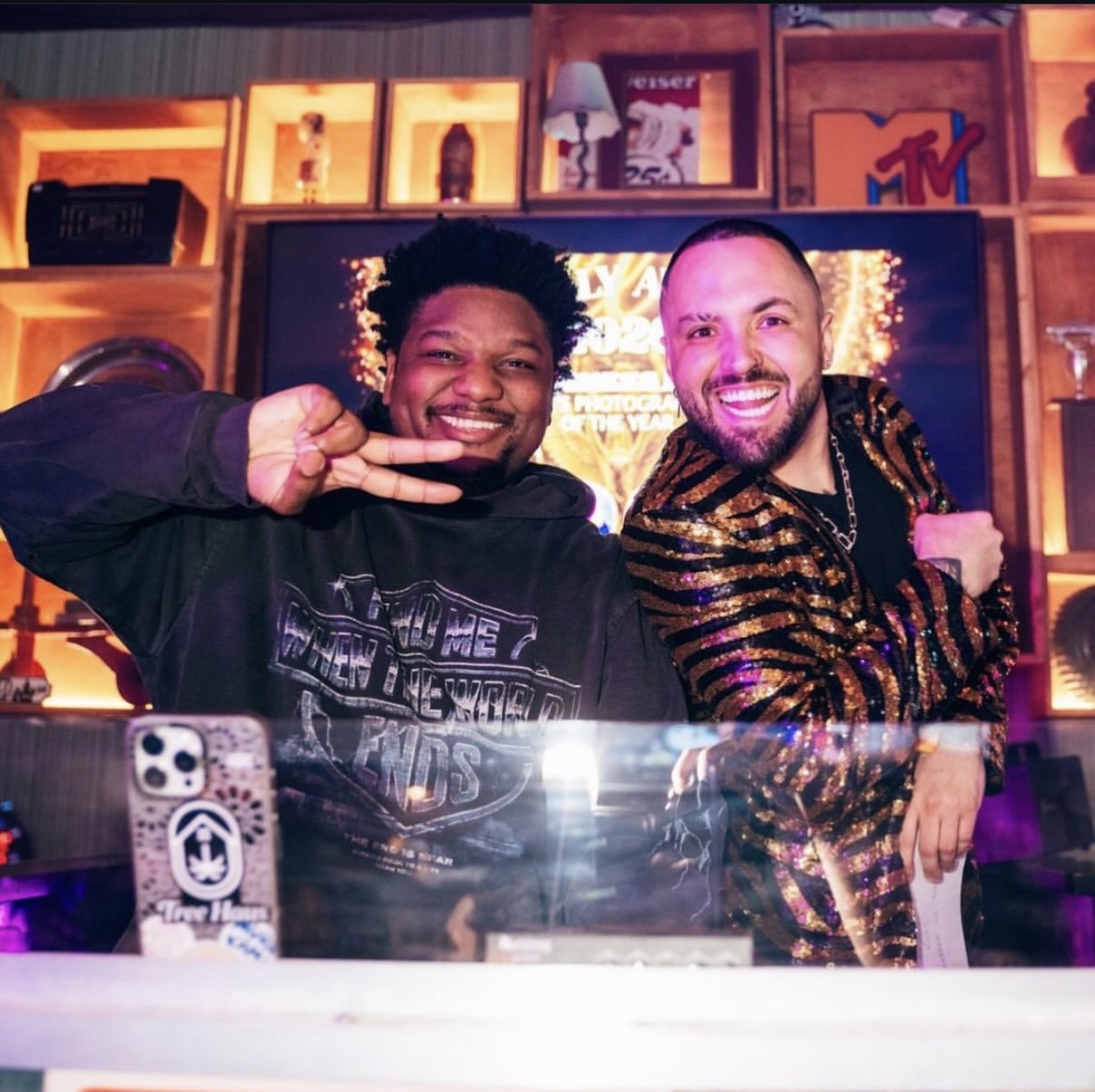 Two smiling men pose together in a colorful room with shelves and TV behind them.