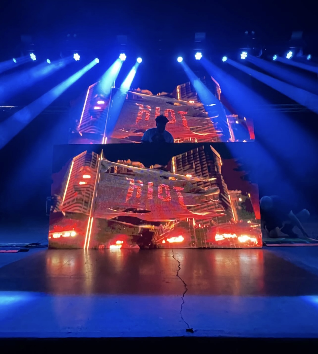 Stage with DJ booth illuminated by blue and red lights, person standing behind the DJ equipment, large screen displaying visuals.