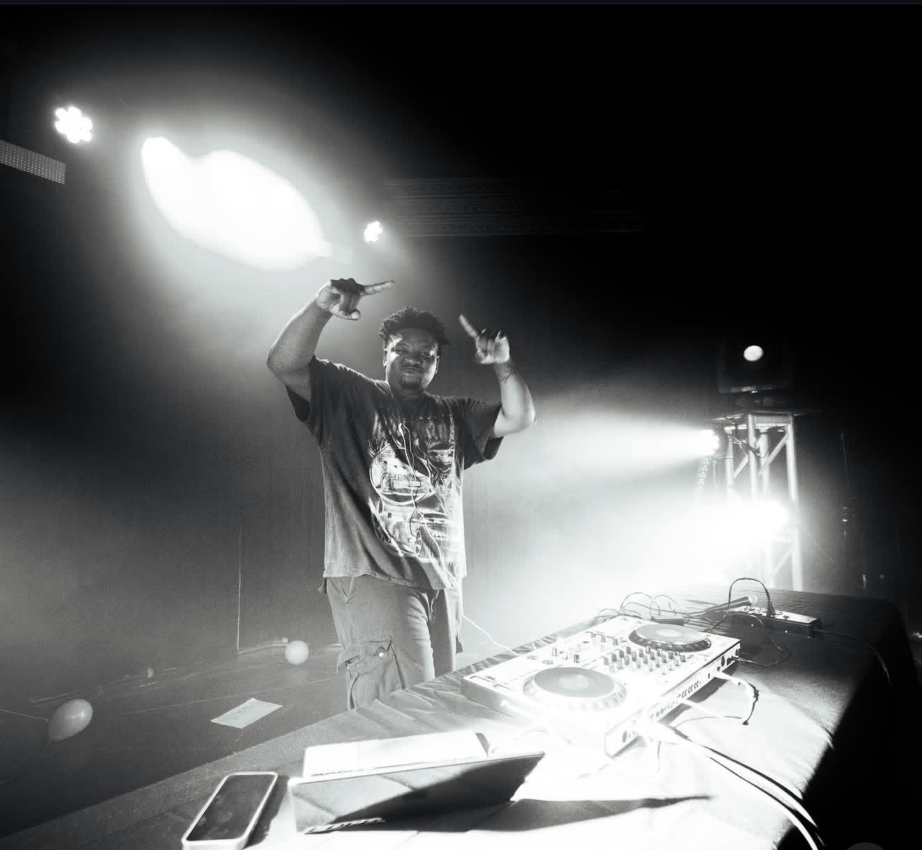 DJ standing behind a mixing console on stage, raising both hands, with bright stage lights behind him.