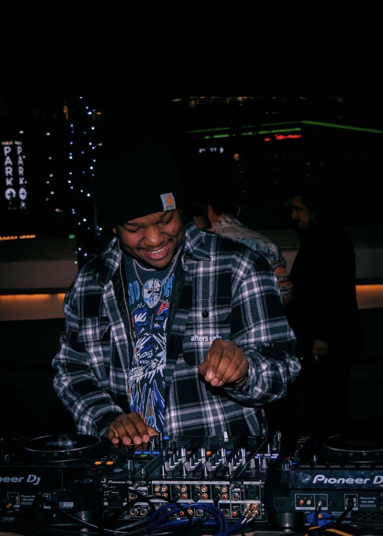Person smiling and dancing at a DJ setup in a dark environment with other people in the background.
