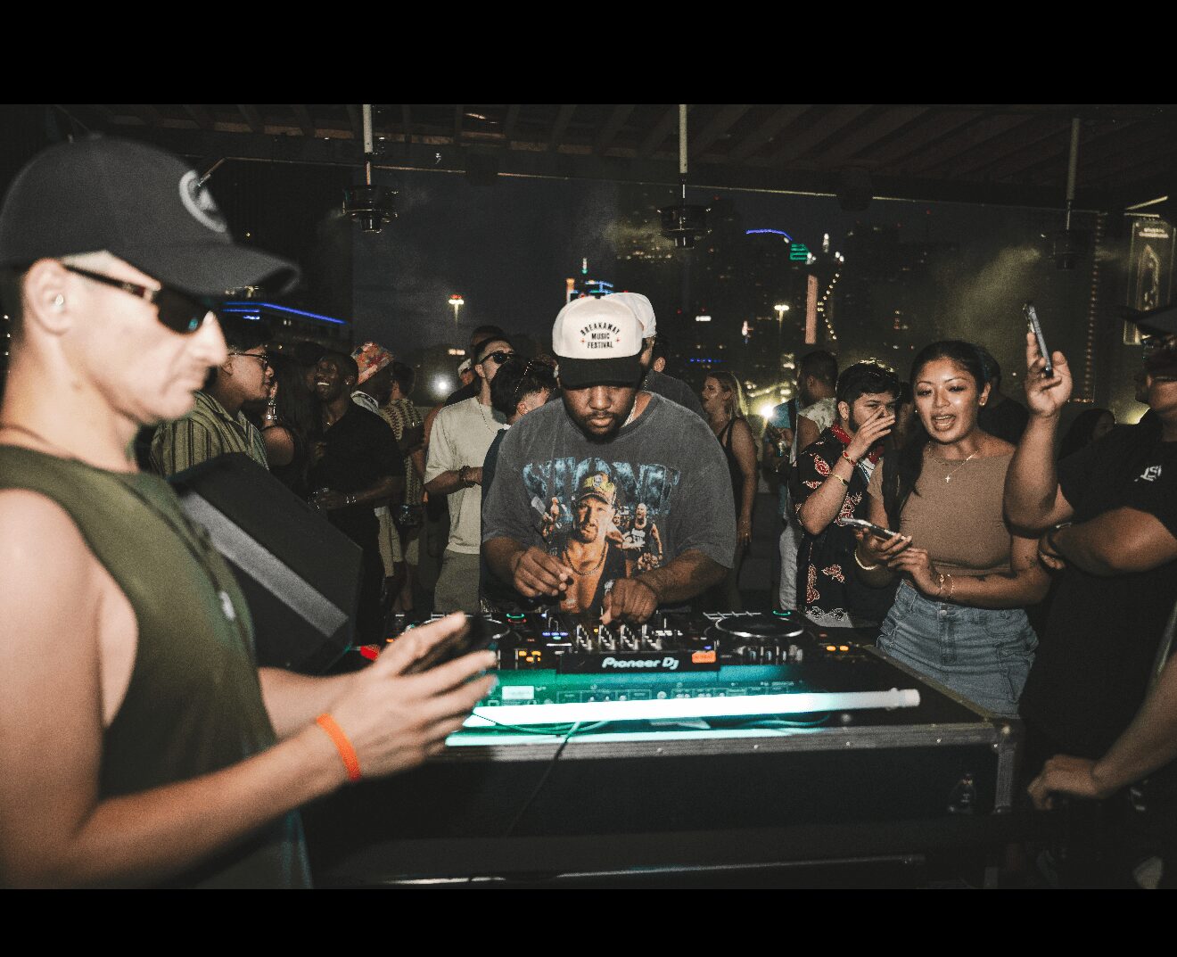 DJ wearing a cap and headphones operates a mixing console in a crowded nightclub.