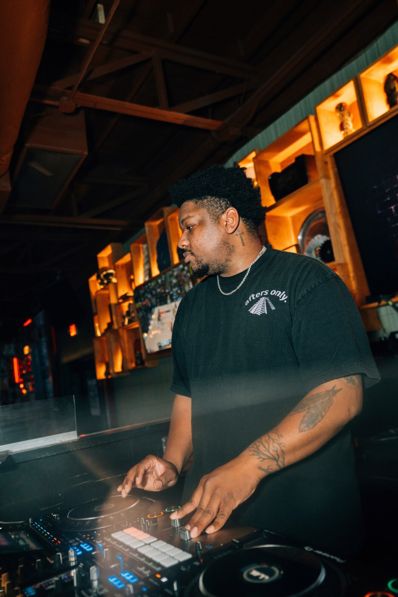 DJ with headphones operating equipment in a dimly lit venue with warm lighting and wooden decor.