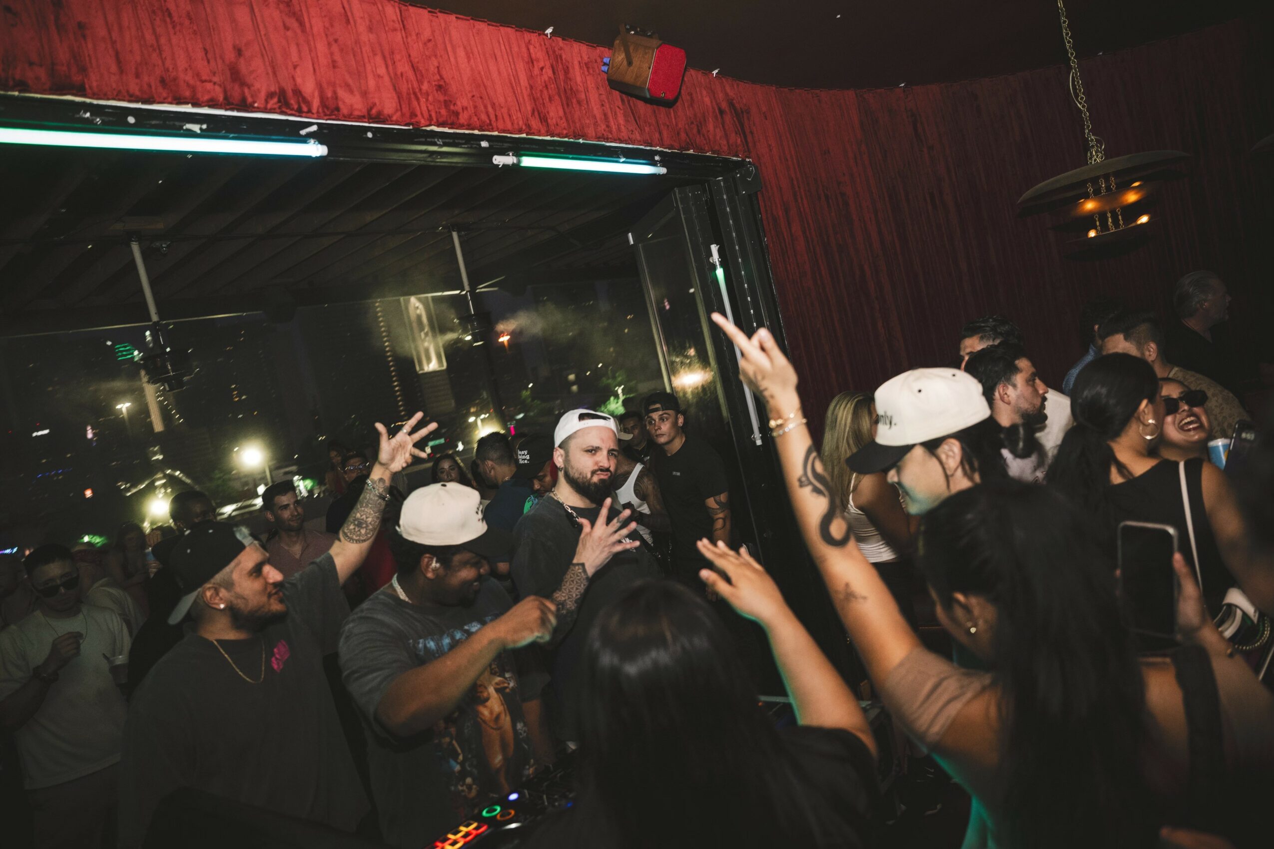 Group of people dancing and raising hands in a dimly lit nightclub with red curtains and reflective windows.