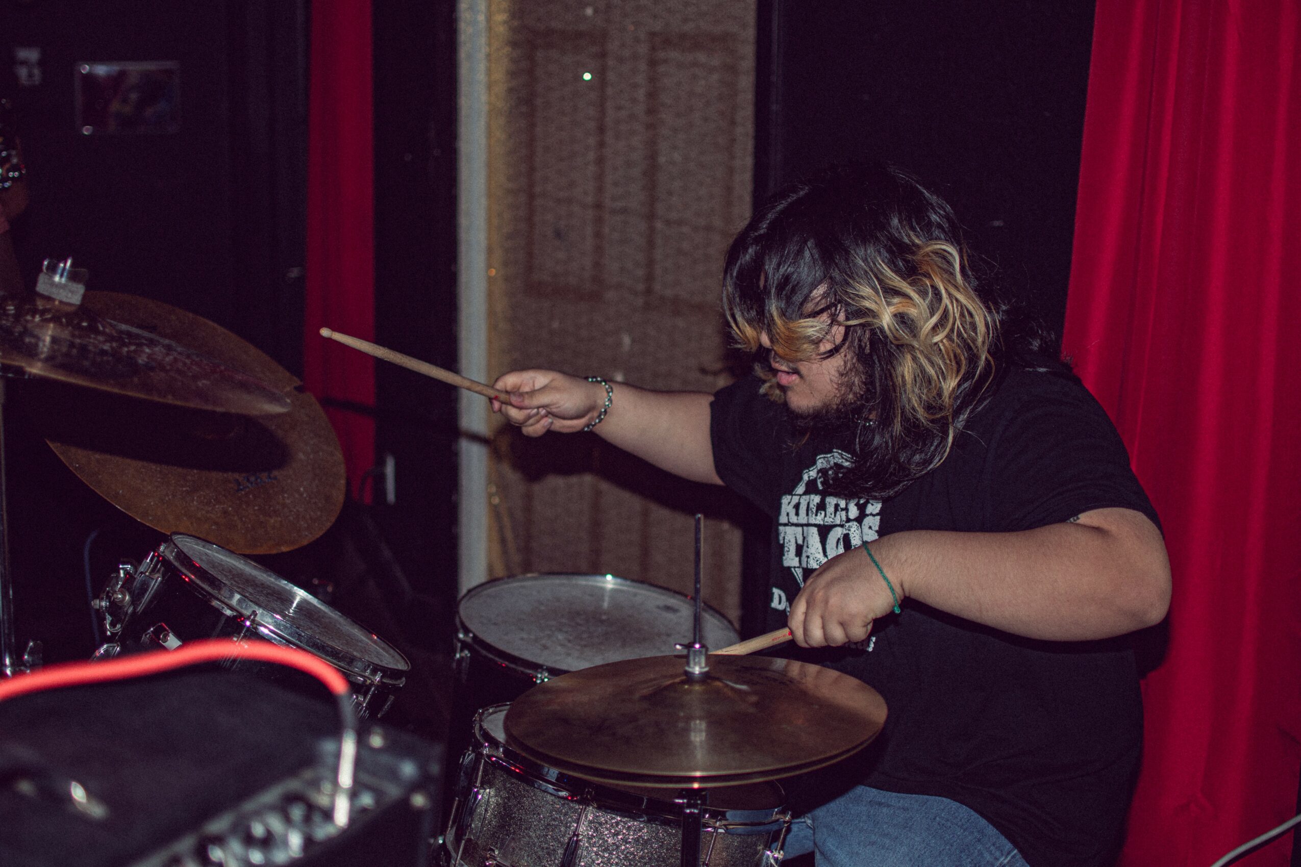Person playing drums with drumsticks, wearing glasses and a black shirt, in a room with red curtains.