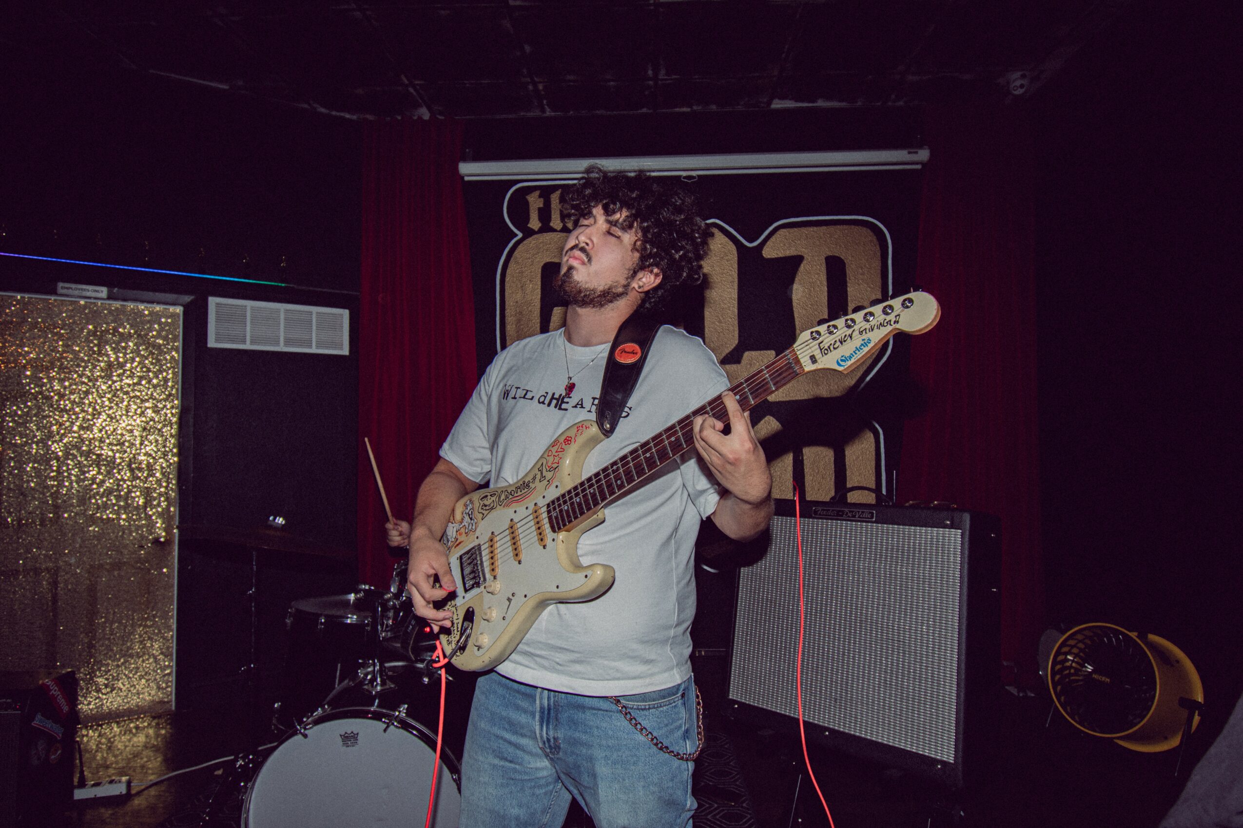 Musician playing electric guitar on stage with drum set and amplifier in background.