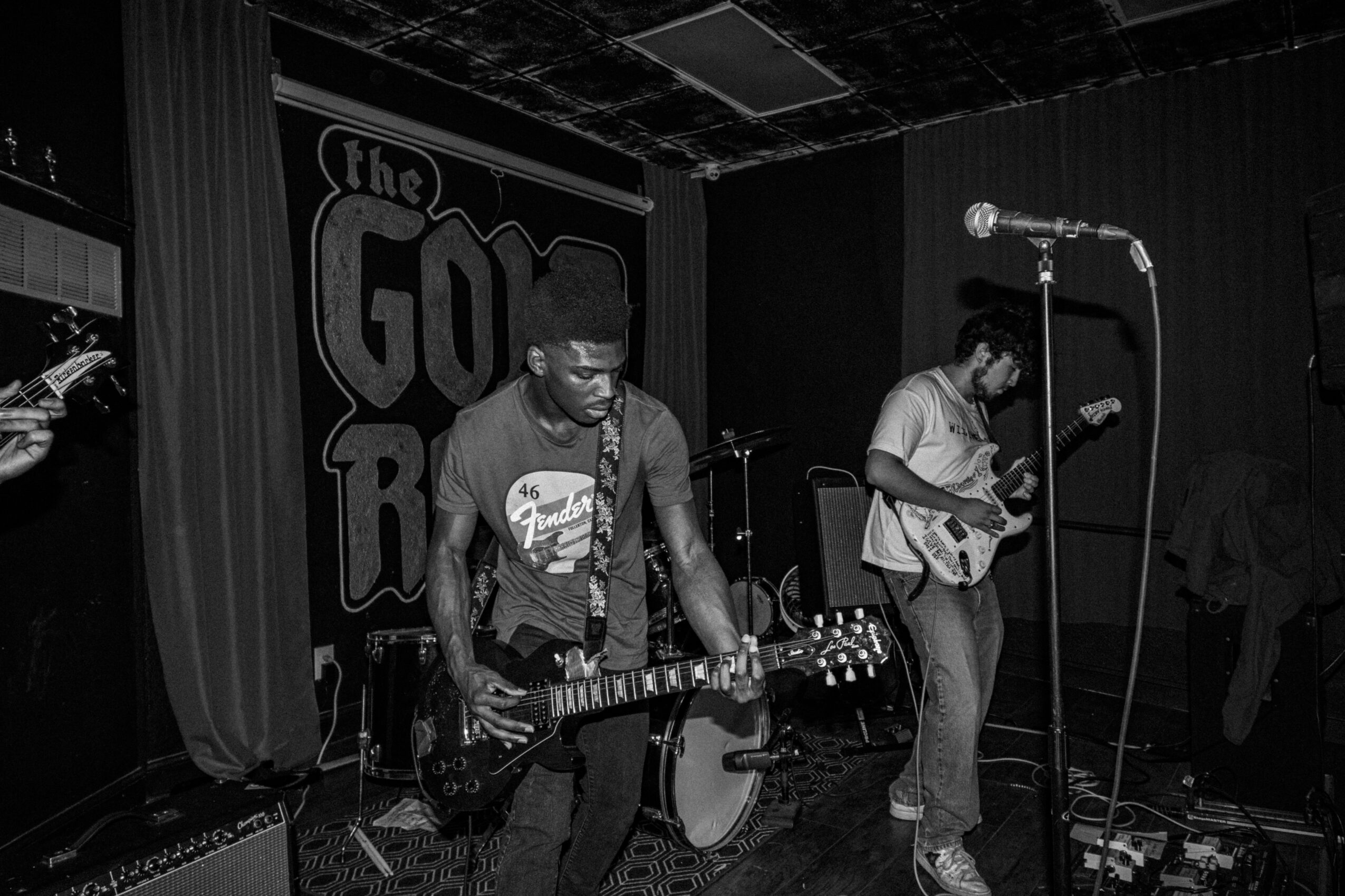 Two musicians perform on stage with a guitar and microphone, in a dimly lit venue with a sign in the background.