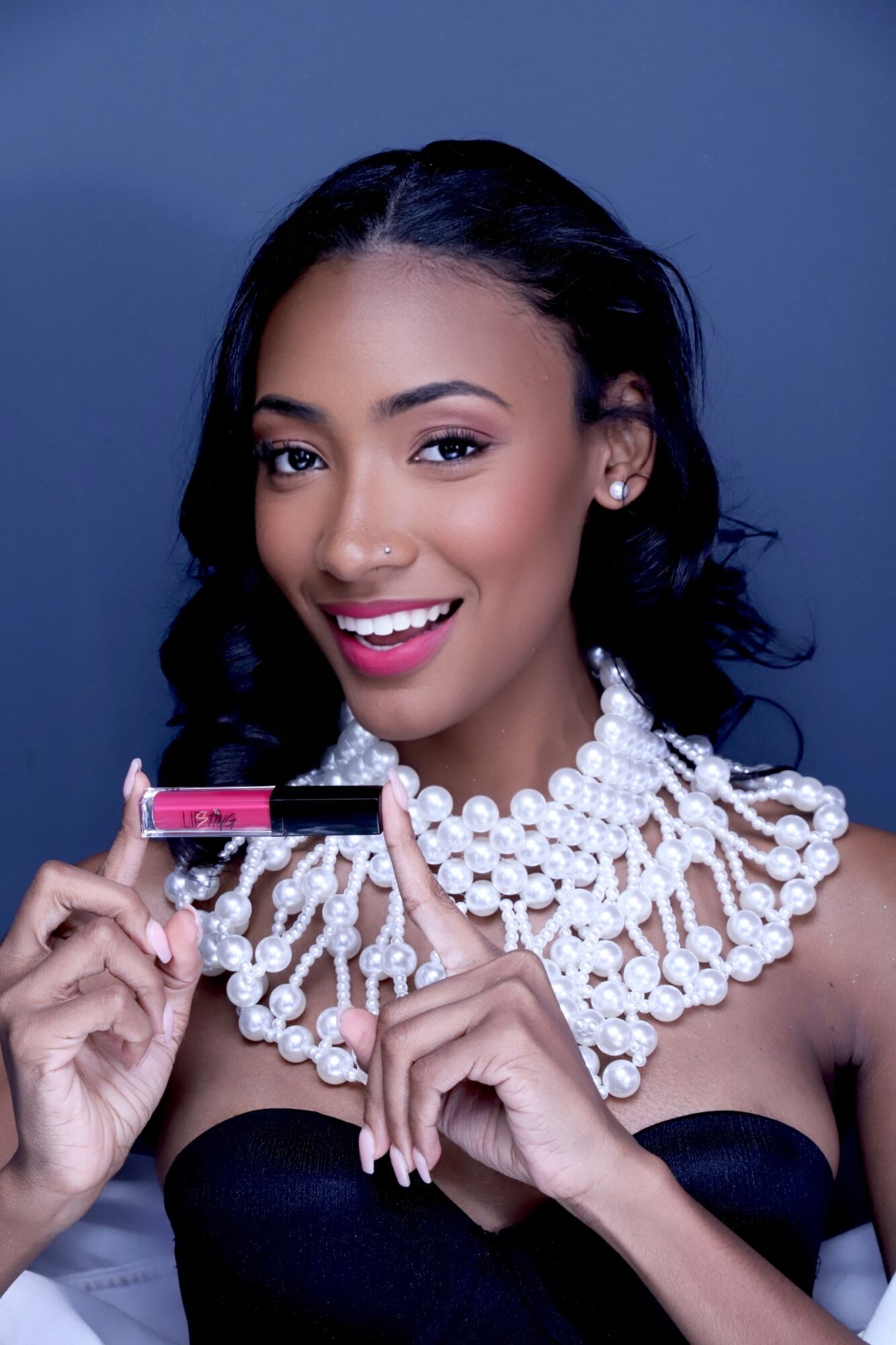 Woman with dark hair, smiling, holding a lipstick, wearing pearl jewelry and a strapless top.