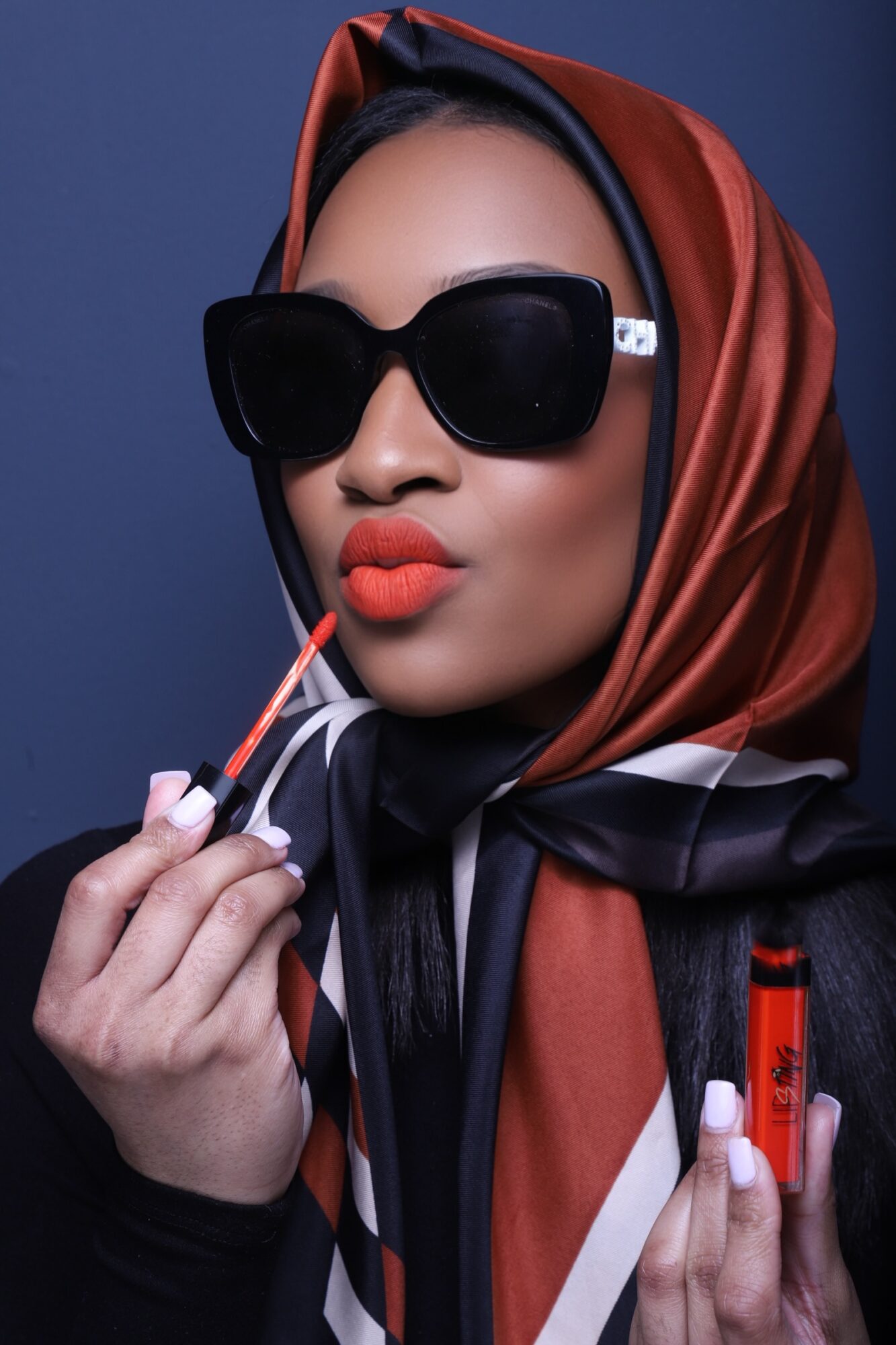 Woman wearing large sunglasses and a colorful headscarf, holding a lipstick and a lip gloss tube.